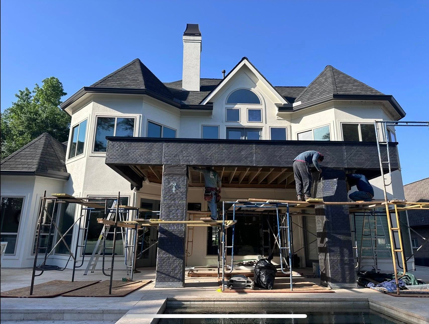 Two workers use scaffolding to install black sheathing on the exterior of a large, light-colored house near a pool.