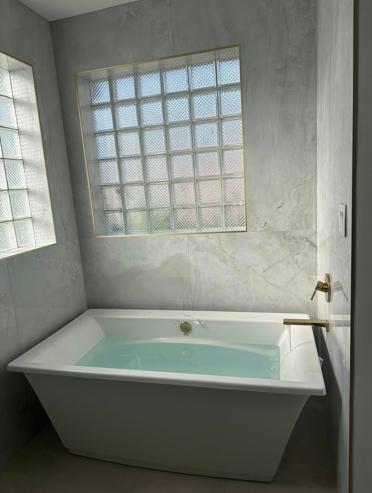 A white, rectangular freestanding bathtub filled with water, positioned in a corner with textured gray walls and glass blocks.
