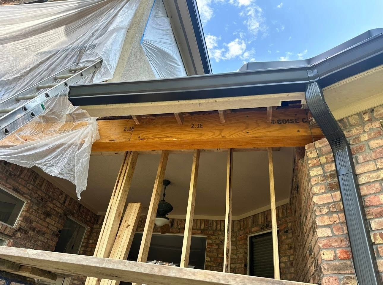A low-angle view of a home exterior under renovation, showing an exposed wooden beam, wall studs, and new dark gutters.