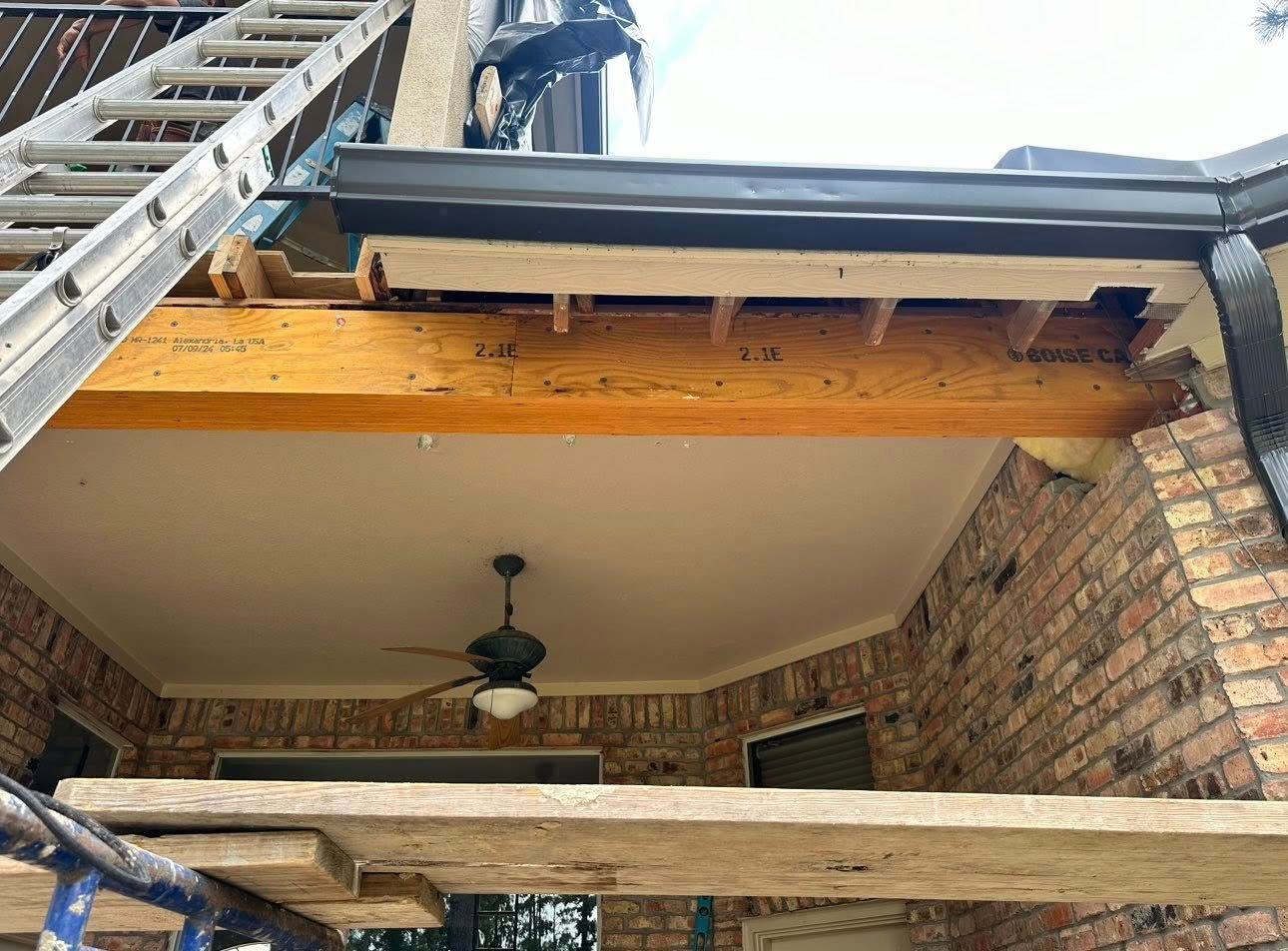 A construction site shows a large wooden beam supporting an outdoor roof overhang with a metal ladder and scaffolding nearby.