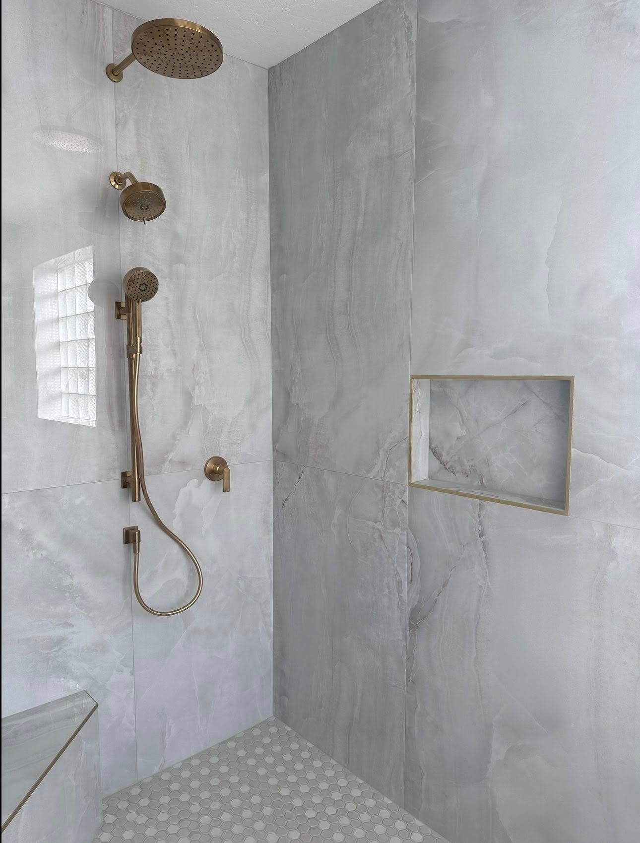 Modern walk-in shower with light gray marble walls, a bronze showerhead and handheld sprayer, and a recessed wall niche.