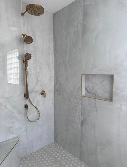 A shower stall with large, light-grey marble-patterned tiles, bronze fixtures, a recessed shelf, and a mosaic tiled floor.