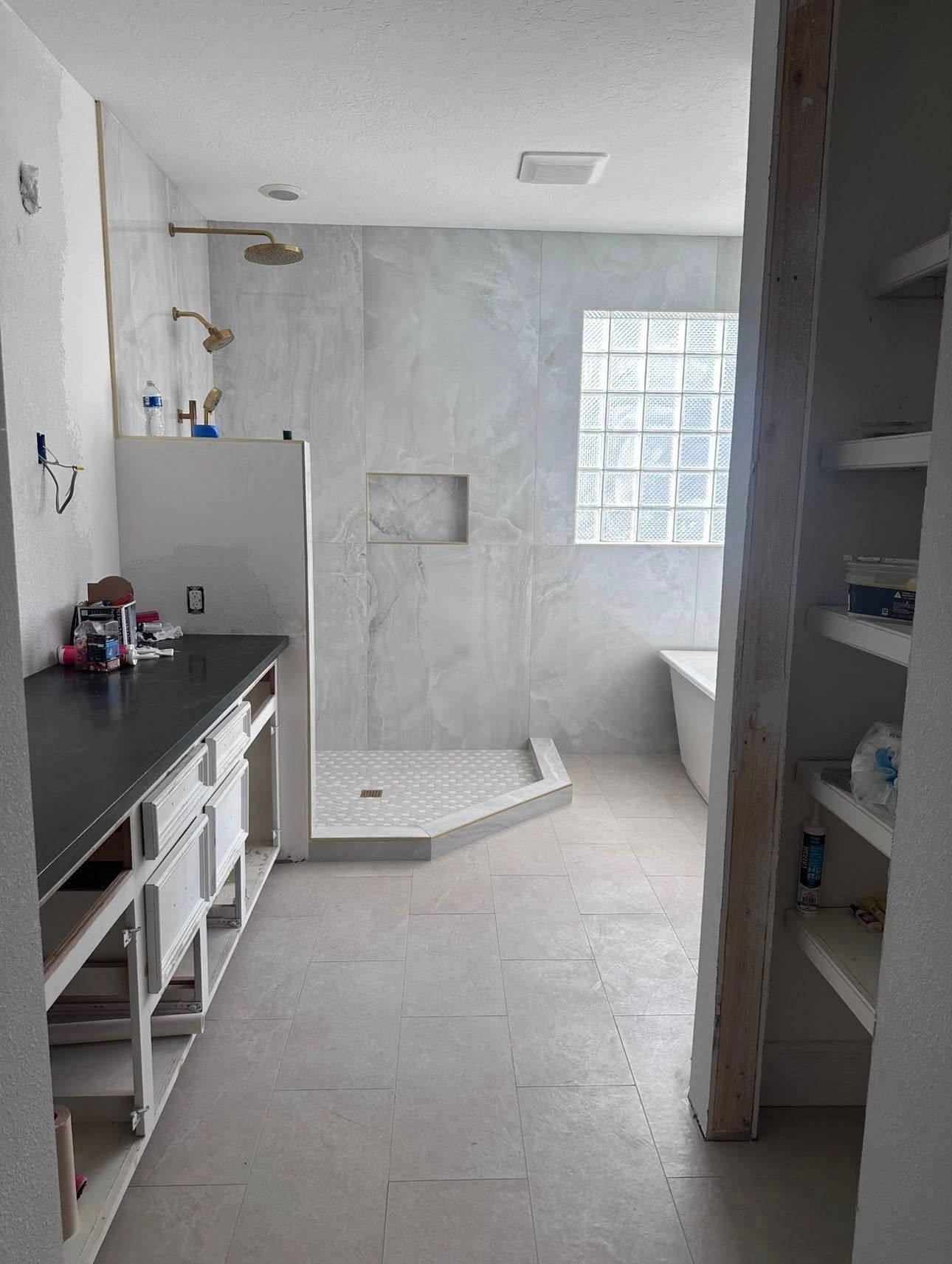 A light gray bathroom featuring a walk-in shower with a glass block window, a dark-topped vanity, and open wall shelving.