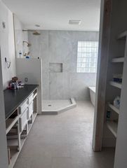 A spacious bathroom with a dark vanity, a walk-in shower with large gray tiles, a bathtub, and open wooden shelving.
