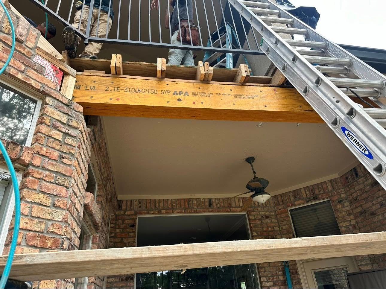 Contractors work on a residential balcony renovation, featuring a wooden support beam and a metal ladder against brick walls.