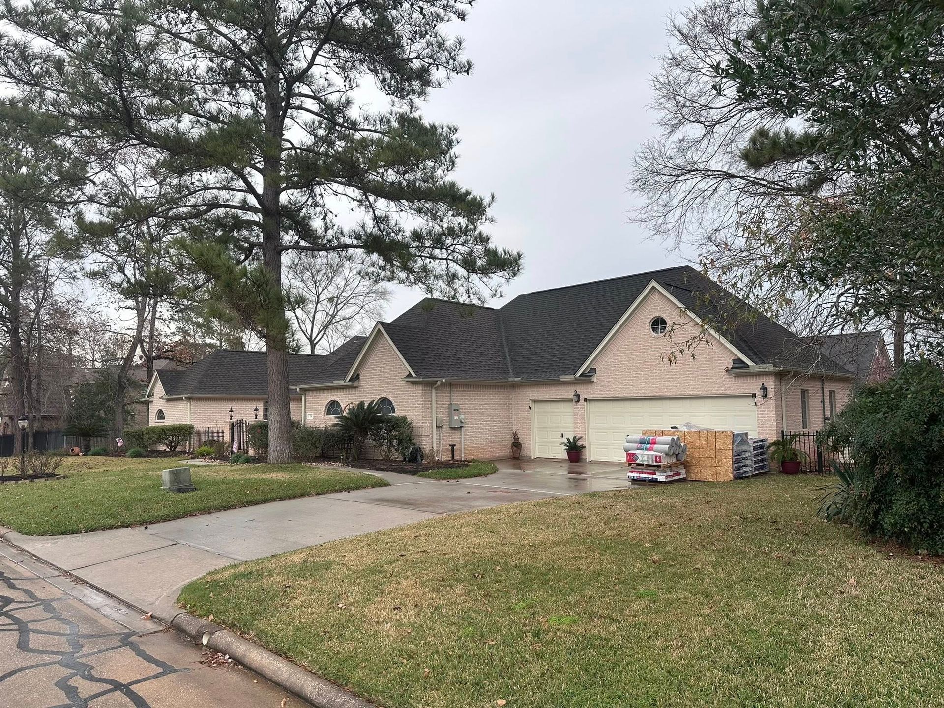 A single-story, light-brick suburban house with a dark shingled roof, a paved driveway, and trees under an overcast sky.