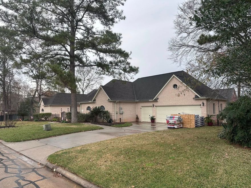 A single-story, light-brick suburban house with a dark gabled roof, a three-car garage, and a large lawn under cloudy skies.
