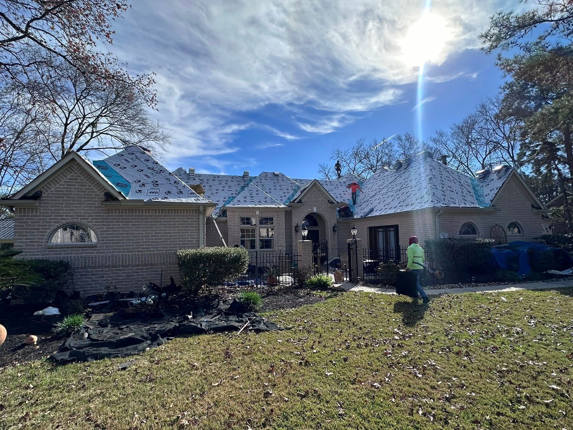 Workers install a new roof on a light-colored brick house under a sunny blue sky.