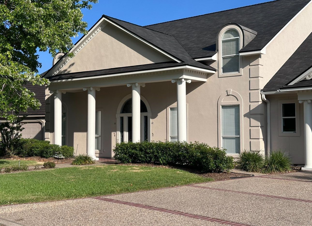 A beige house with a covered front porch featuring four white columns, a black roof, and a gravel driveway.