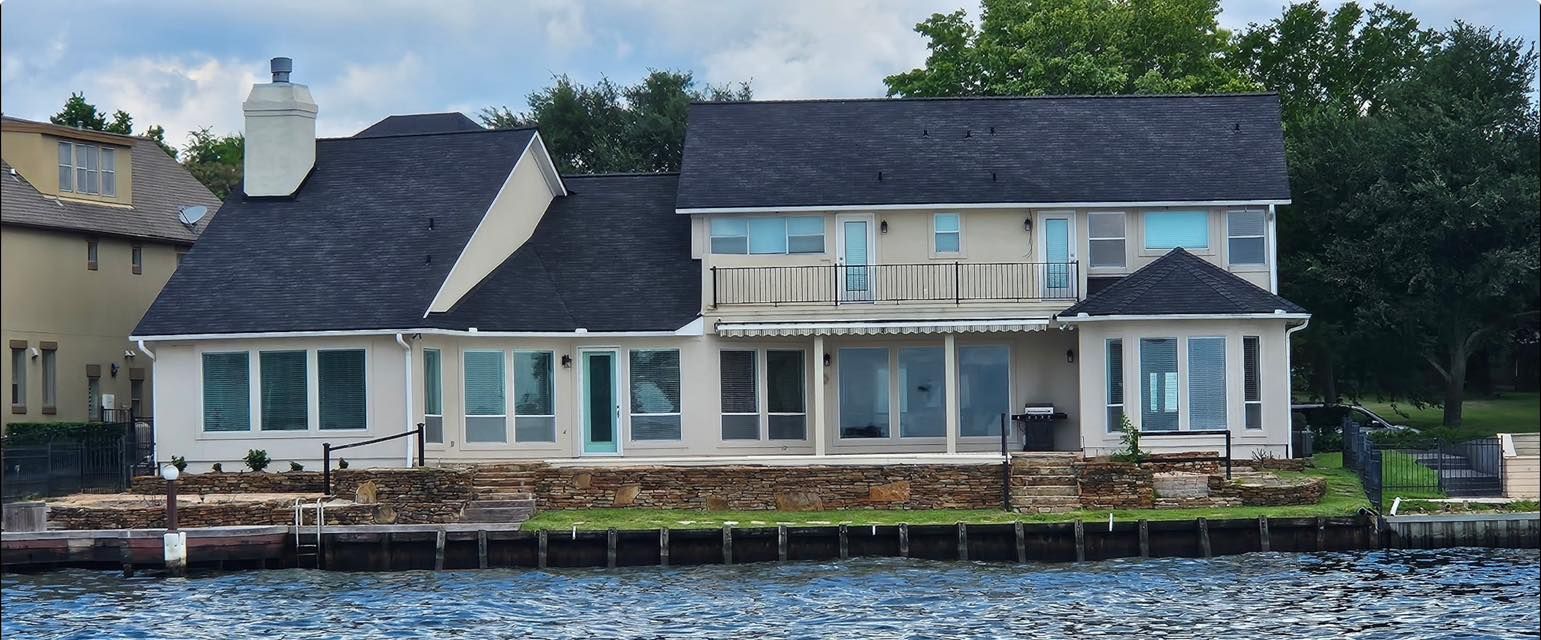 A large, two-story house with white siding and a black roof sits along a lakefront with a stone retaining wall.