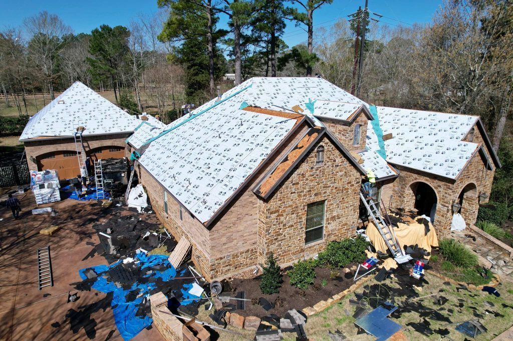 An aerial view shows a brick house undergoing a roof replacement, with white underlayment installed on the roof.