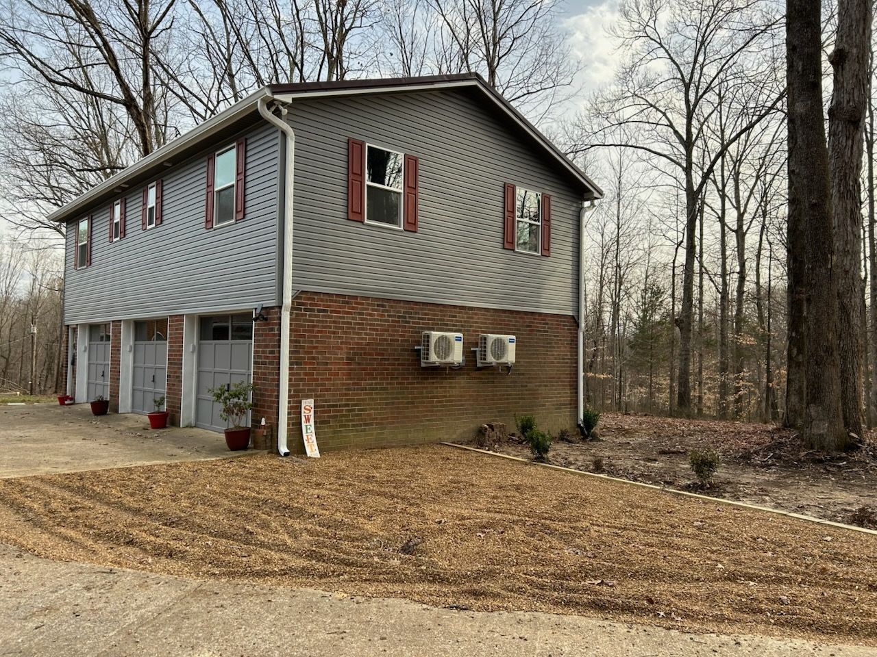 Two-story gray house with three garage doors, red shutters, and brick facade in a wooded area.