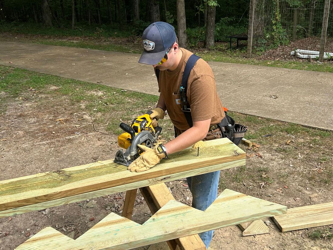 Person using a circular saw to cut a wooden plank outdoors.  Wears safety glasses, gloves, and a tool belt.