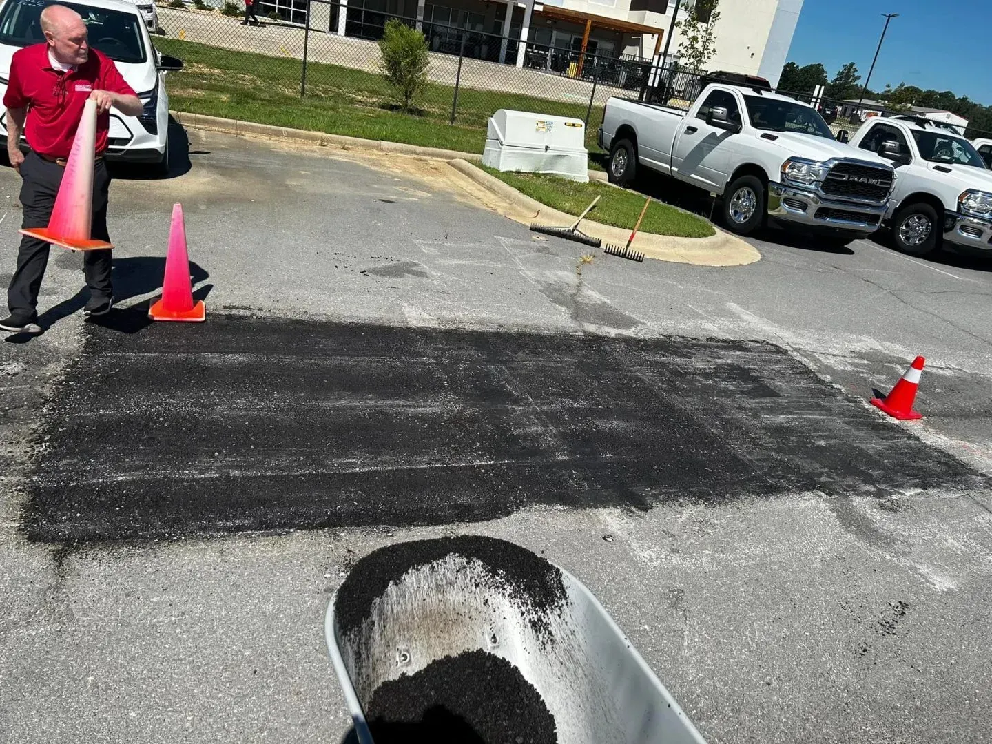 Man pouring asphalt into a parking lot repair, surrounded by traffic cones and work trucks.