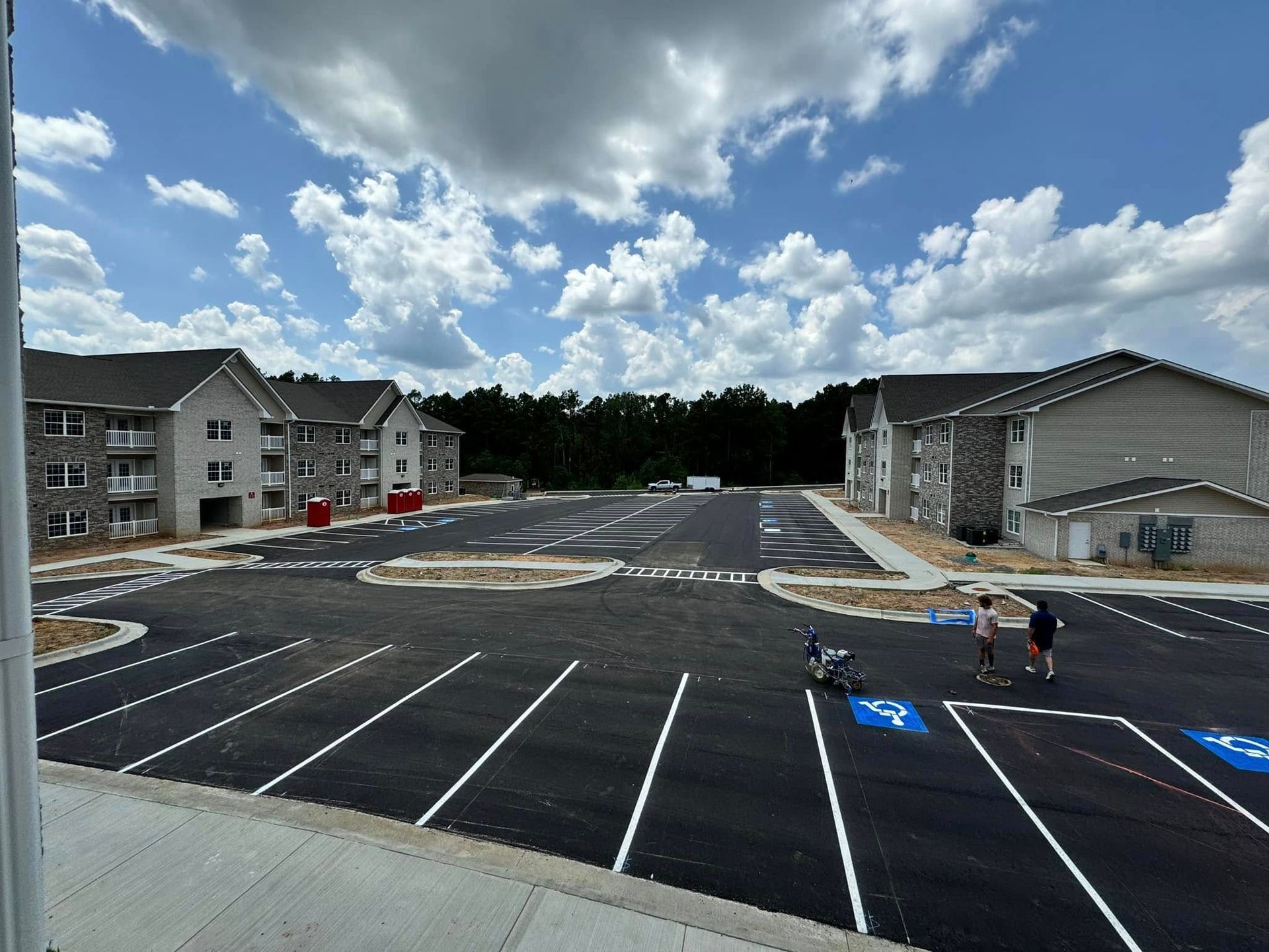 New asphalt parking lot with apartment buildings under cloudy sky. Two people stand near accessible parking.
