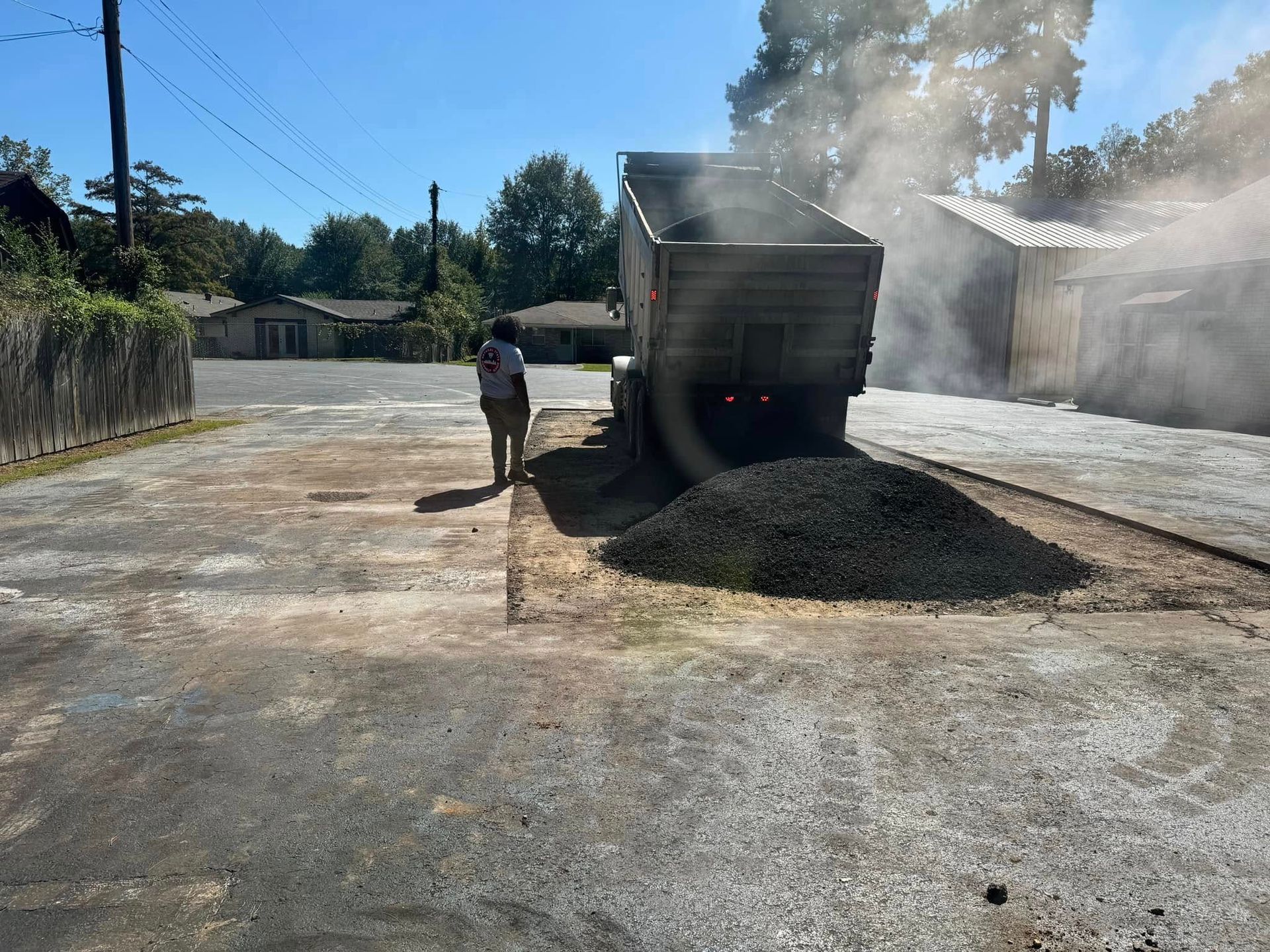 Truck dumping asphalt on a paved road; a person walks near it, with buildings and trees in the background.