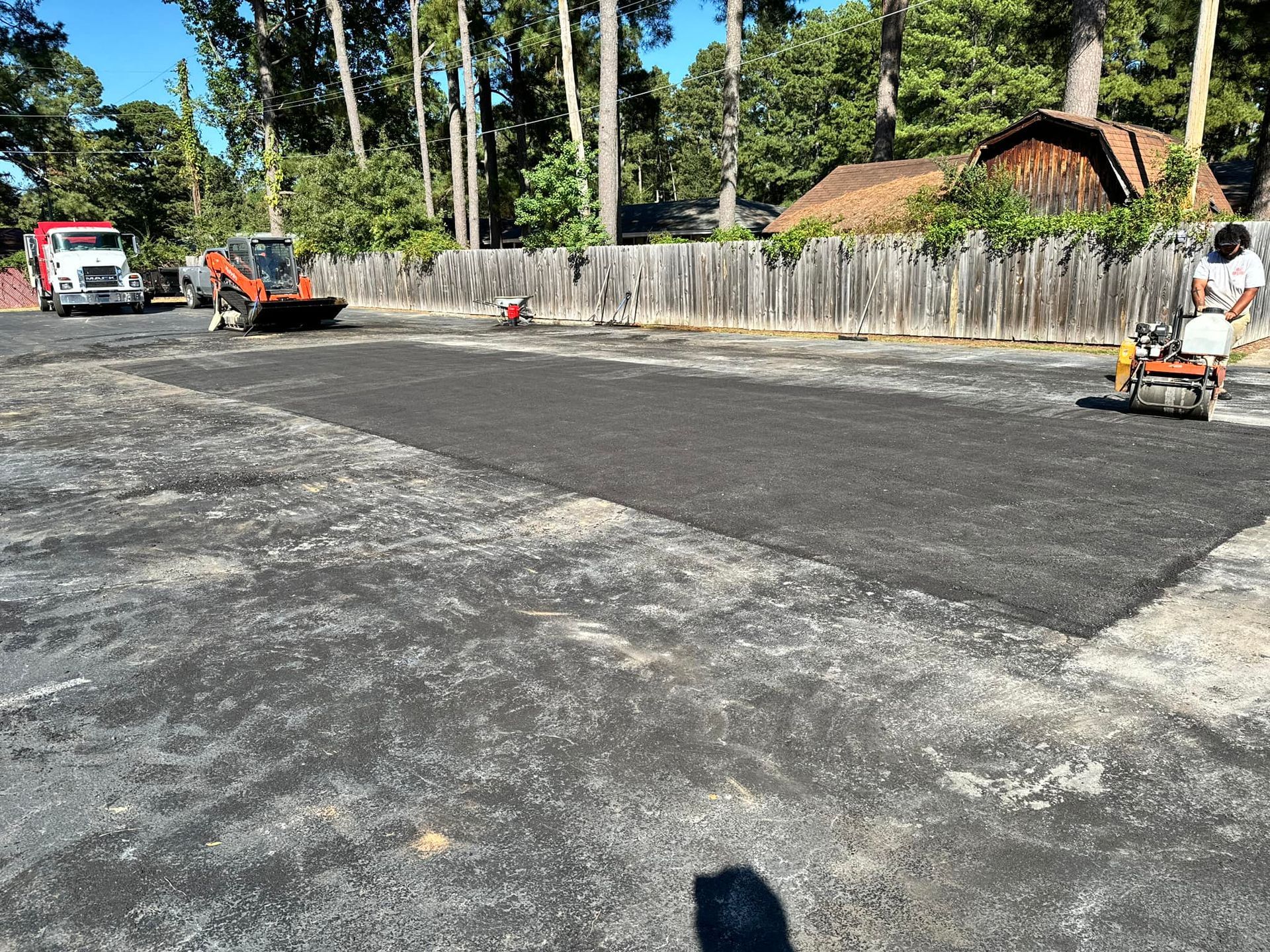 Asphalt paving in progress on a parking lot with a truck, excavator, and worker with a roller.