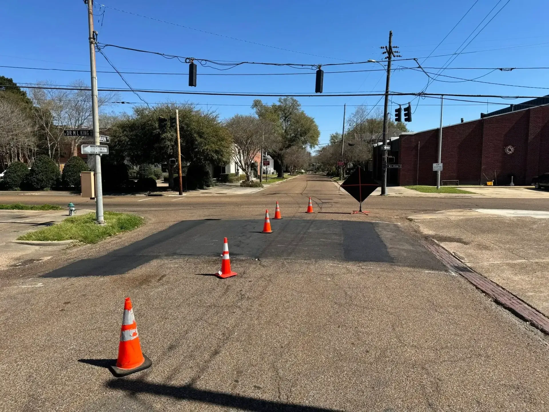 Street with orange traffic cones marking a recent asphalt repair, power lines overhead, and buildings in background.