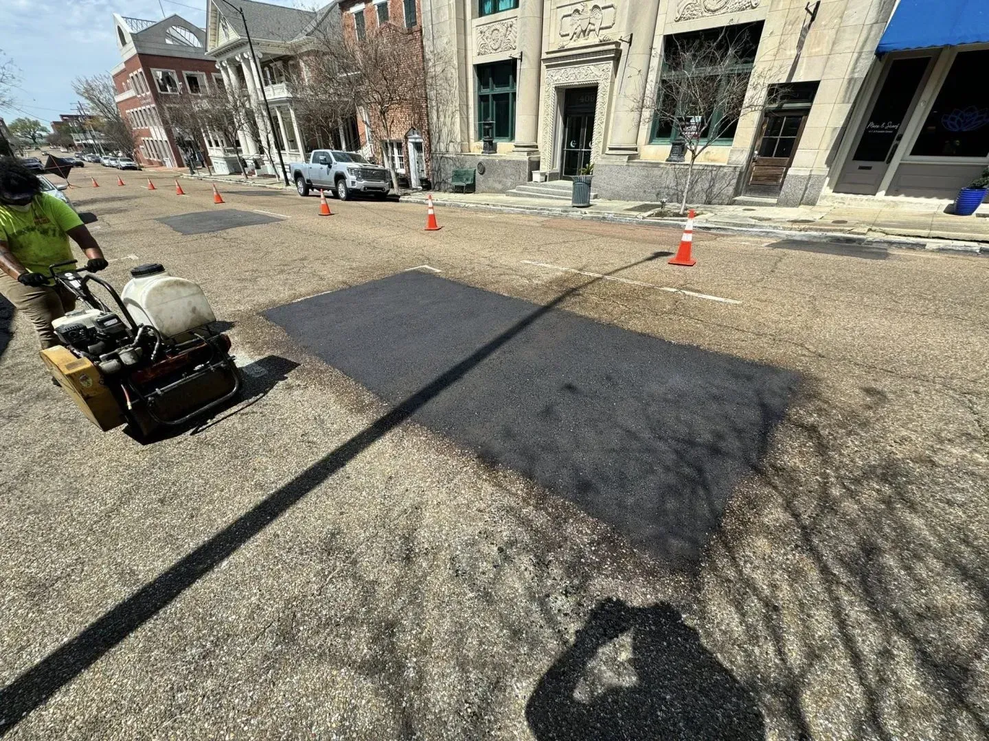 Road resurfacing: Worker using a compactor on a freshly paved area, orange cones, buildings in background.