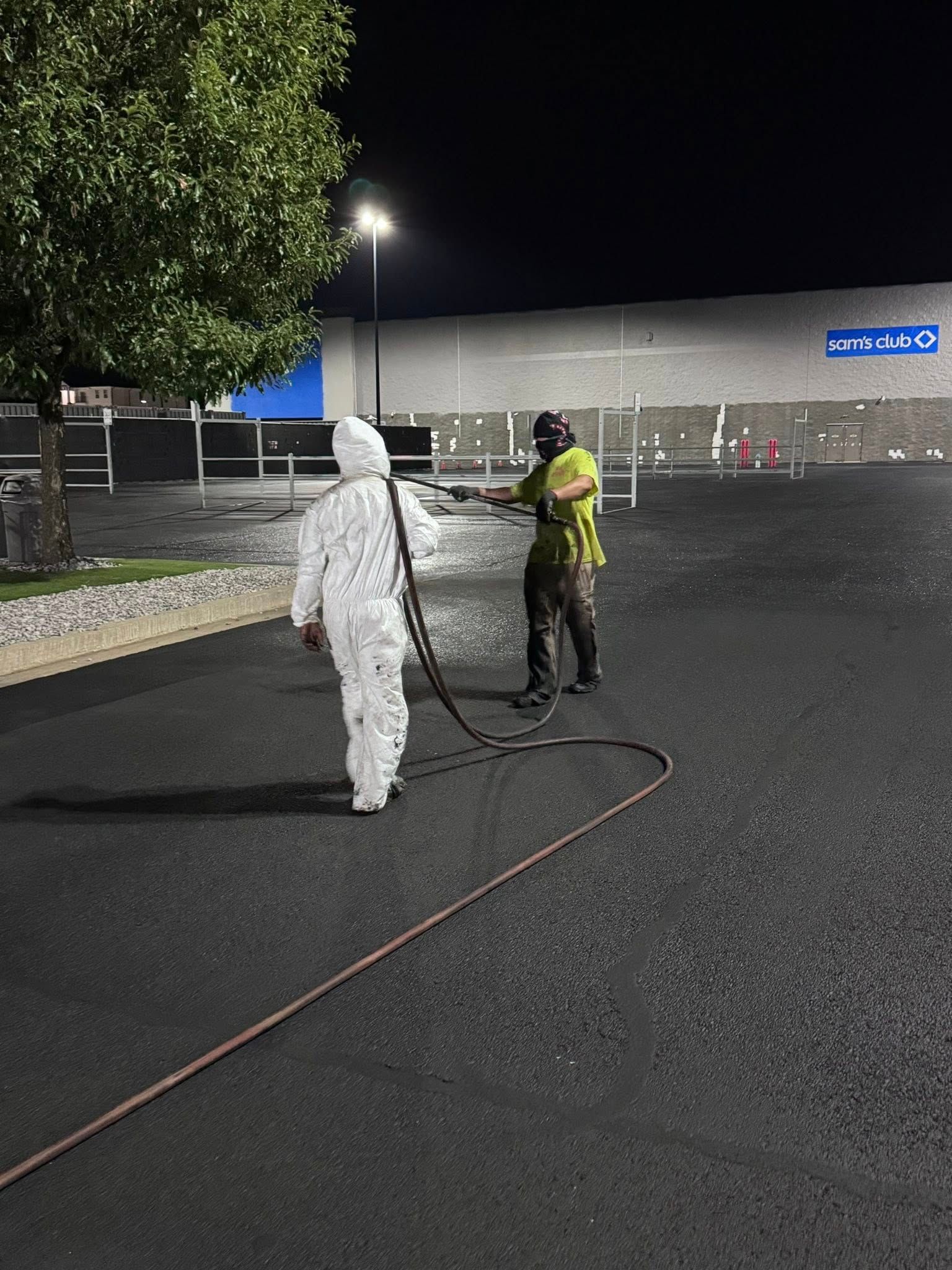 Two people spraying pavement at night near a store. One wears a hazmat suit, the other a vest and mask.