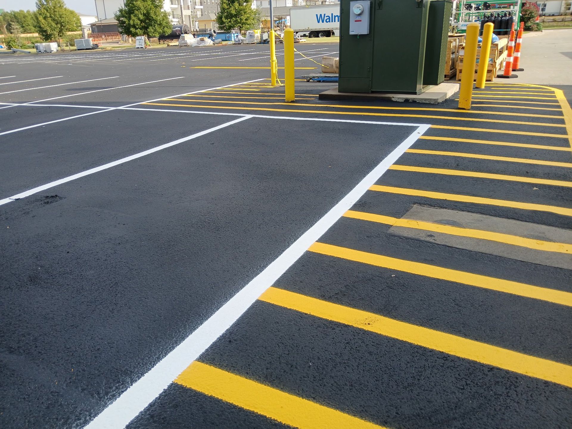 Freshly paved asphalt parking area with yellow and white painted lines, utility box, and bollards.