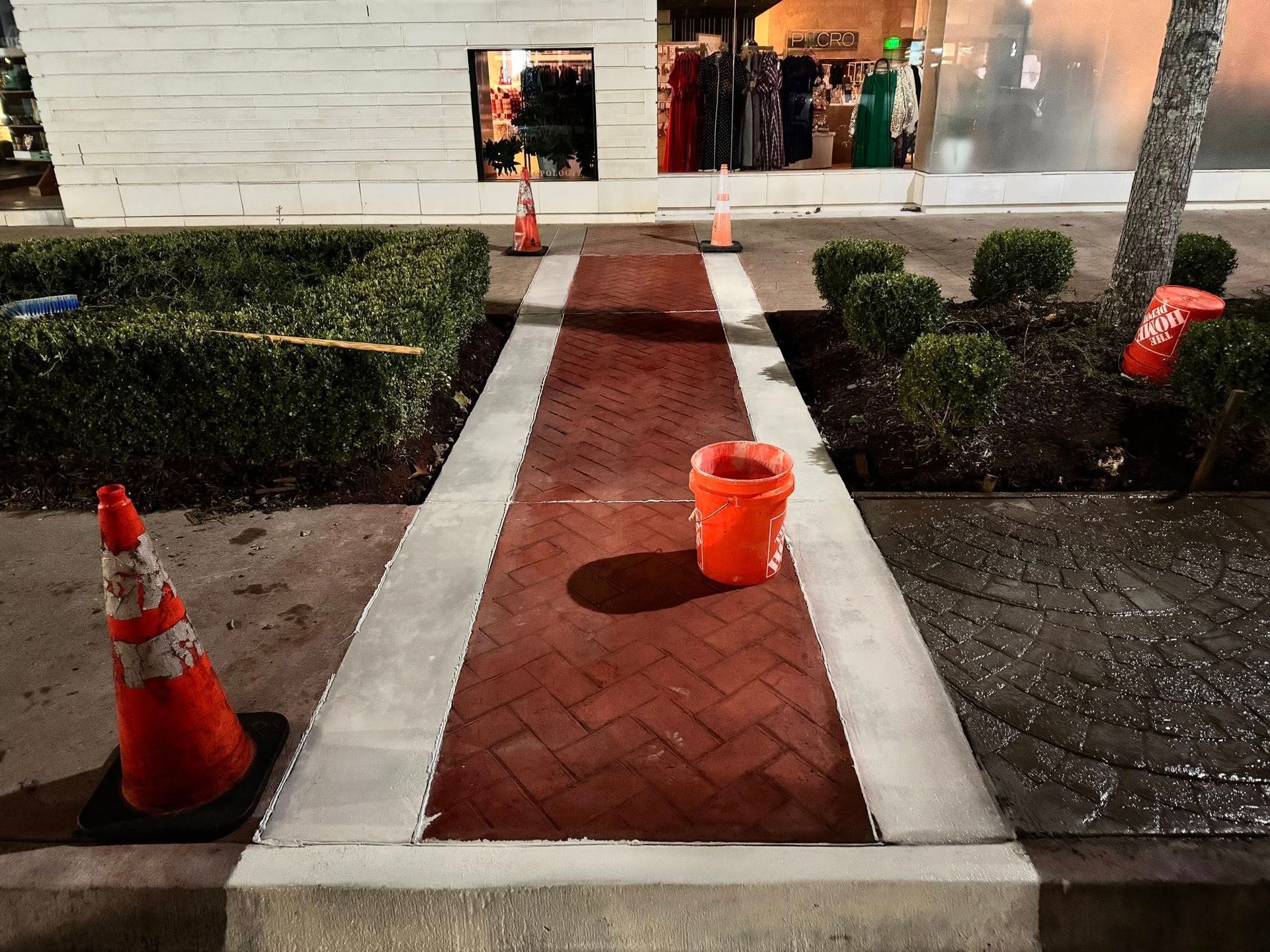 Brick pathway with orange cones and bucket, bordered by concrete and landscaping, in front of a storefront.