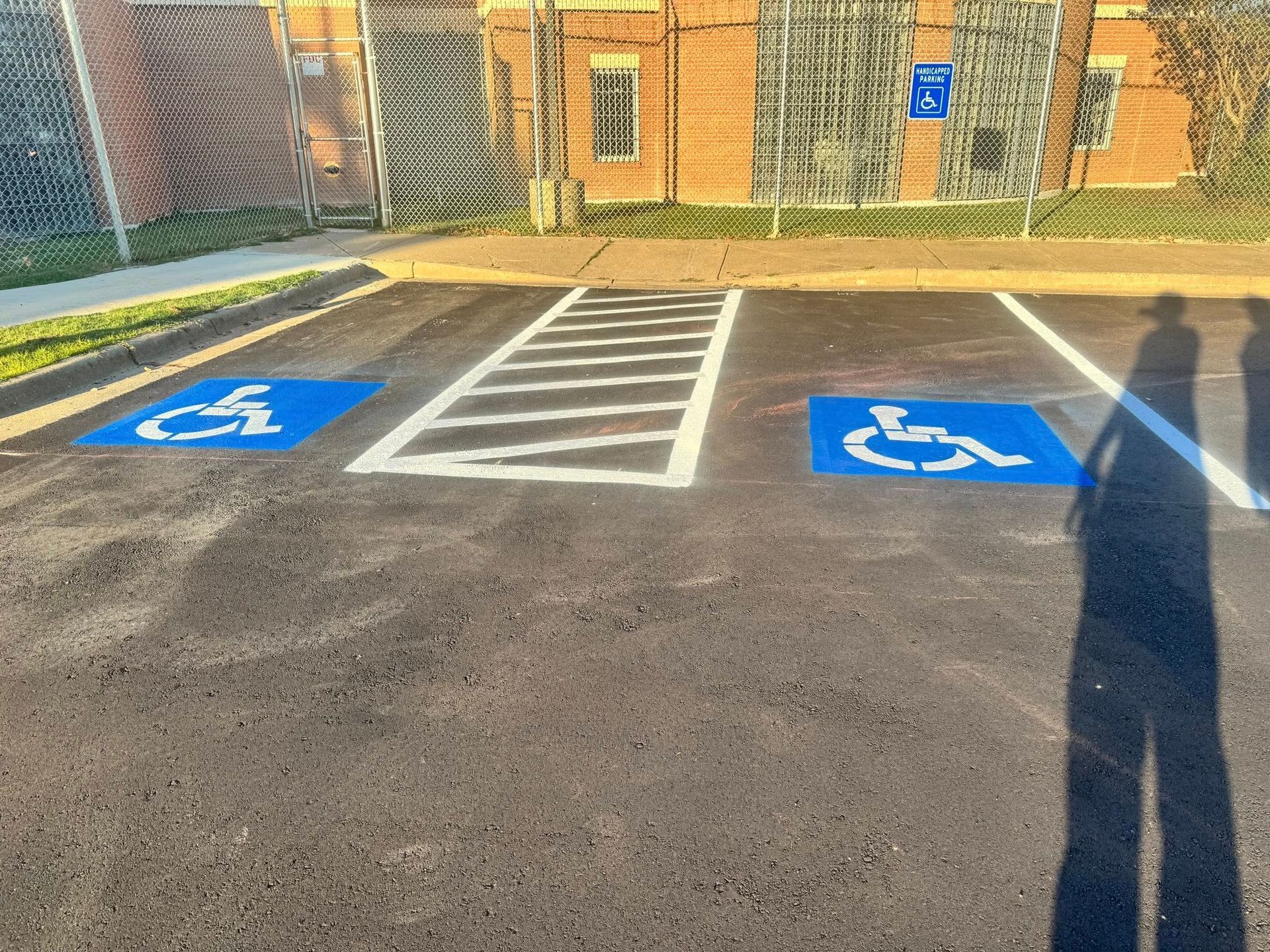 Two blue handicap parking spaces with white striped access area. Shadows of people in the foreground. Brick building in the background.