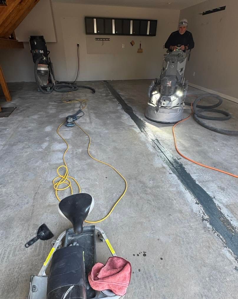 Two workers sand a concrete floor in a garage with hoses, cords, and a floor grinder.