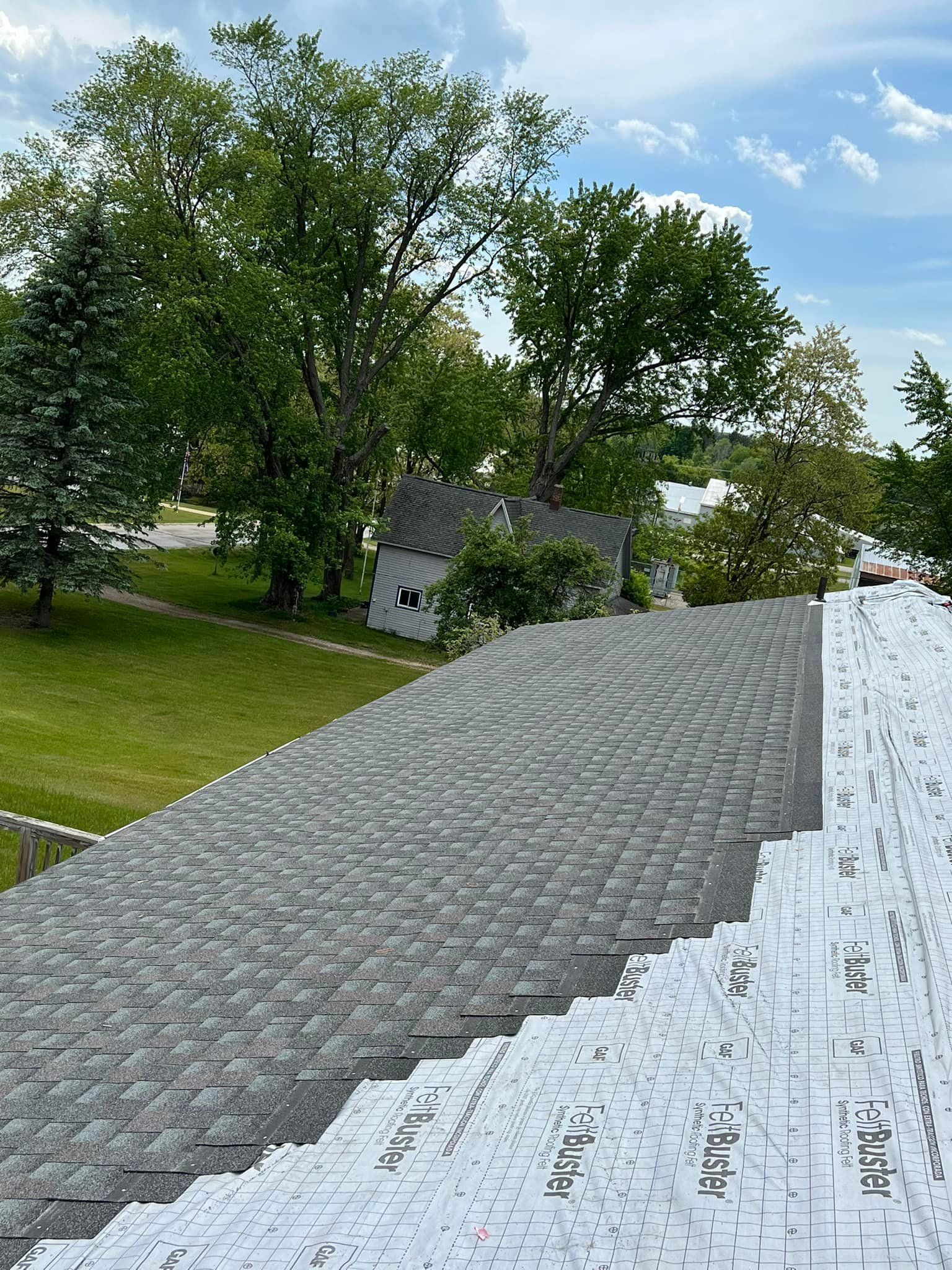 Gray shingle roof, angled view. Distant trees, small building, and blue sky visible.