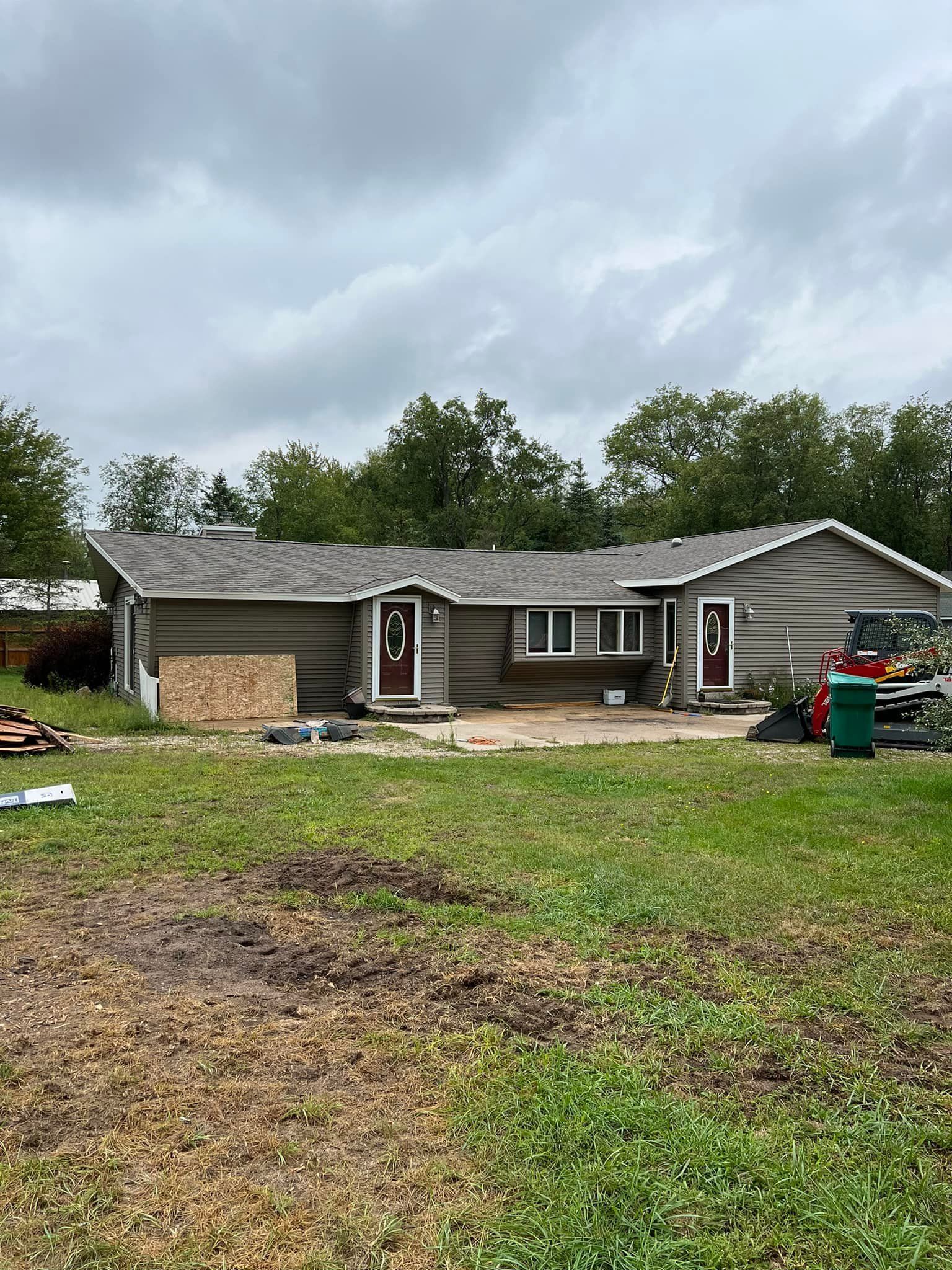 A single-story house with two front doors is under renovation. Exterior is gray; brown doors. Overcast sky.