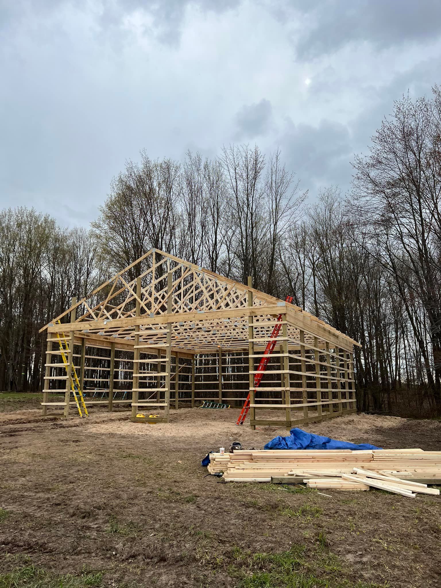 Wooden building frame under construction in a wooded area. Ladders and materials visible. Overcast sky.