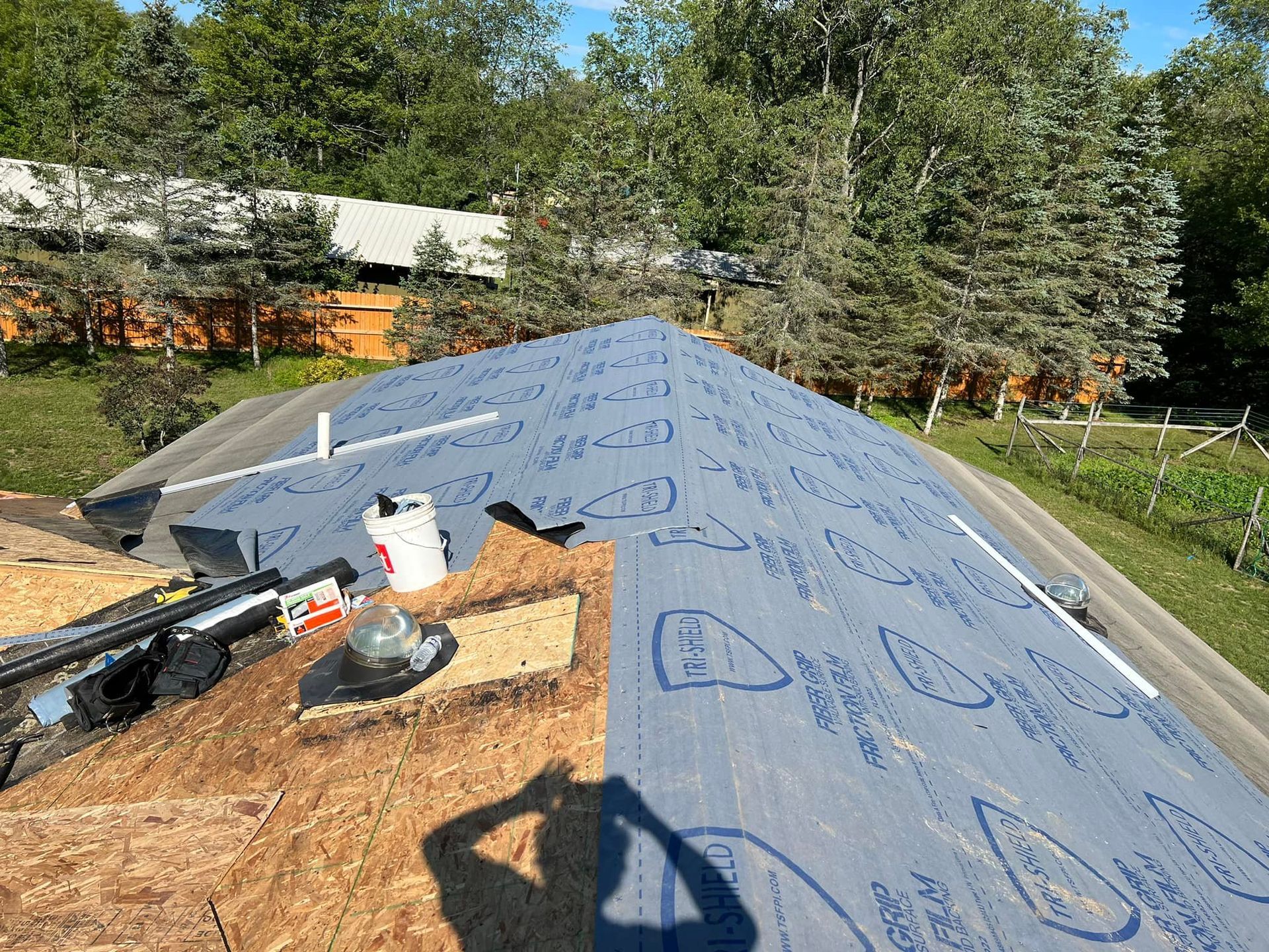 Roof partially covered with blue underlayment and plywood. Tools and materials on the roof.