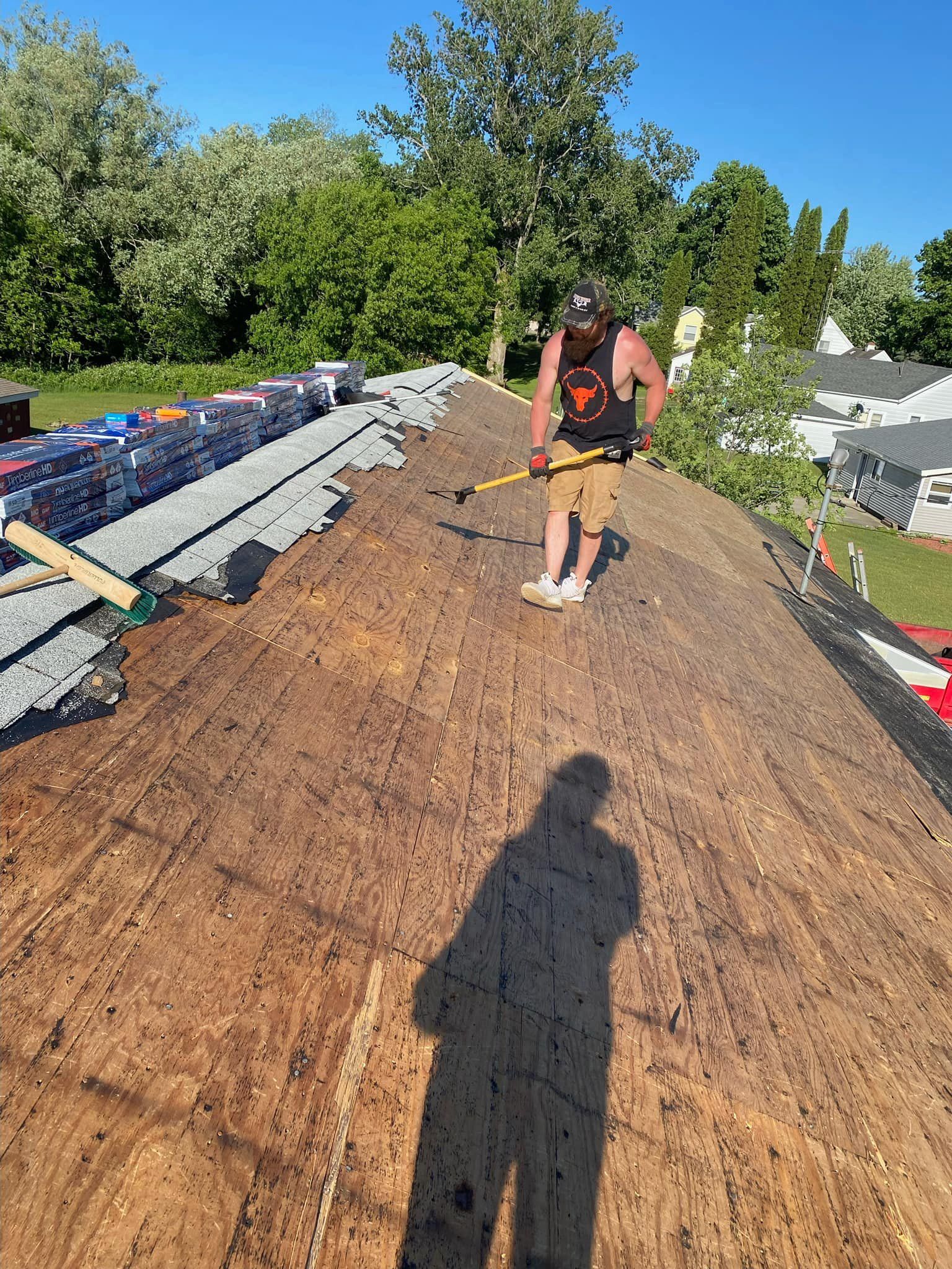 Man on a rooftop, preparing shingles. Sunlight. Green trees in the background. Shadow of the person is visible.