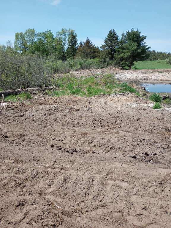 Brown dirt pile in foreground, grassy area and trees by water, blue sky.