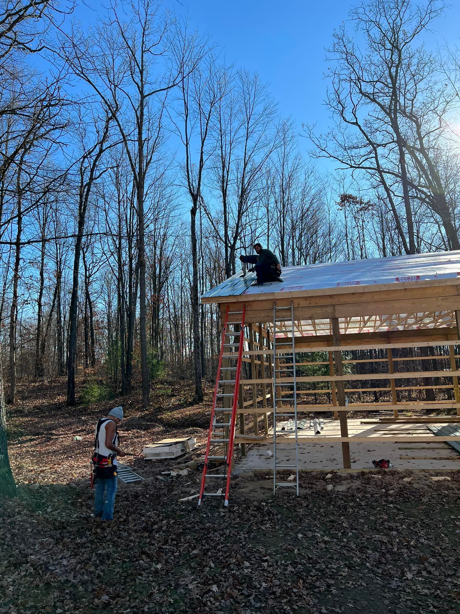 Two people building a wooden structure in a wooded area; one on the roof, one on the ground.