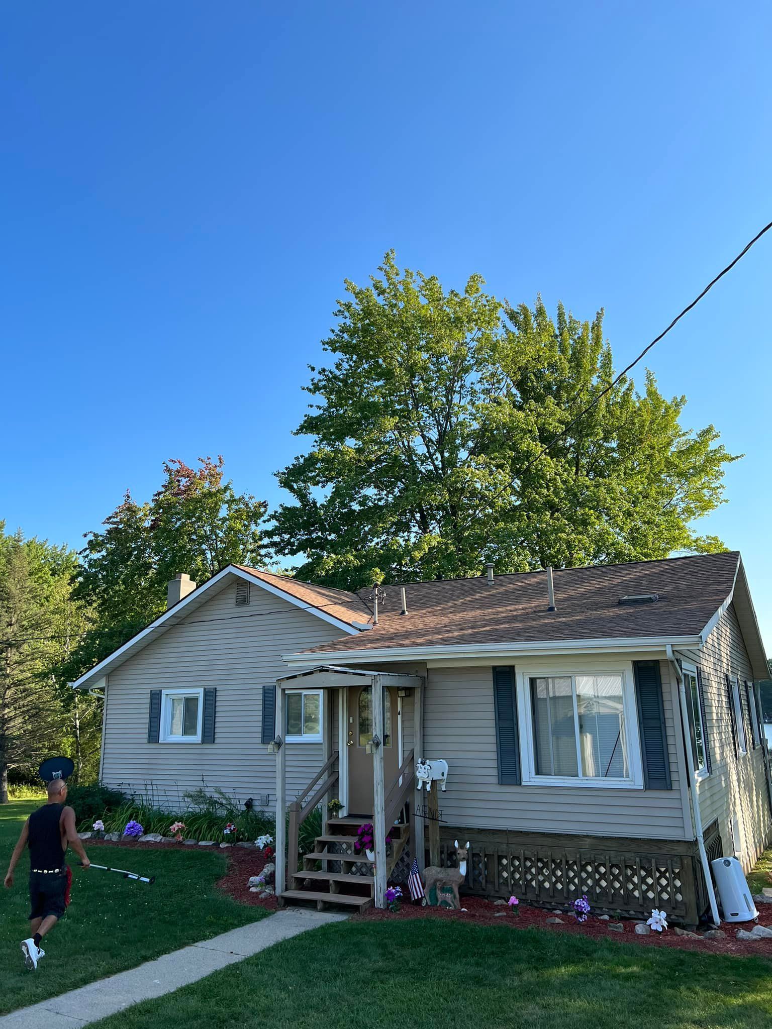 Small, light-colored house with a dark roof. A person walks by in the yard under a clear blue sky.