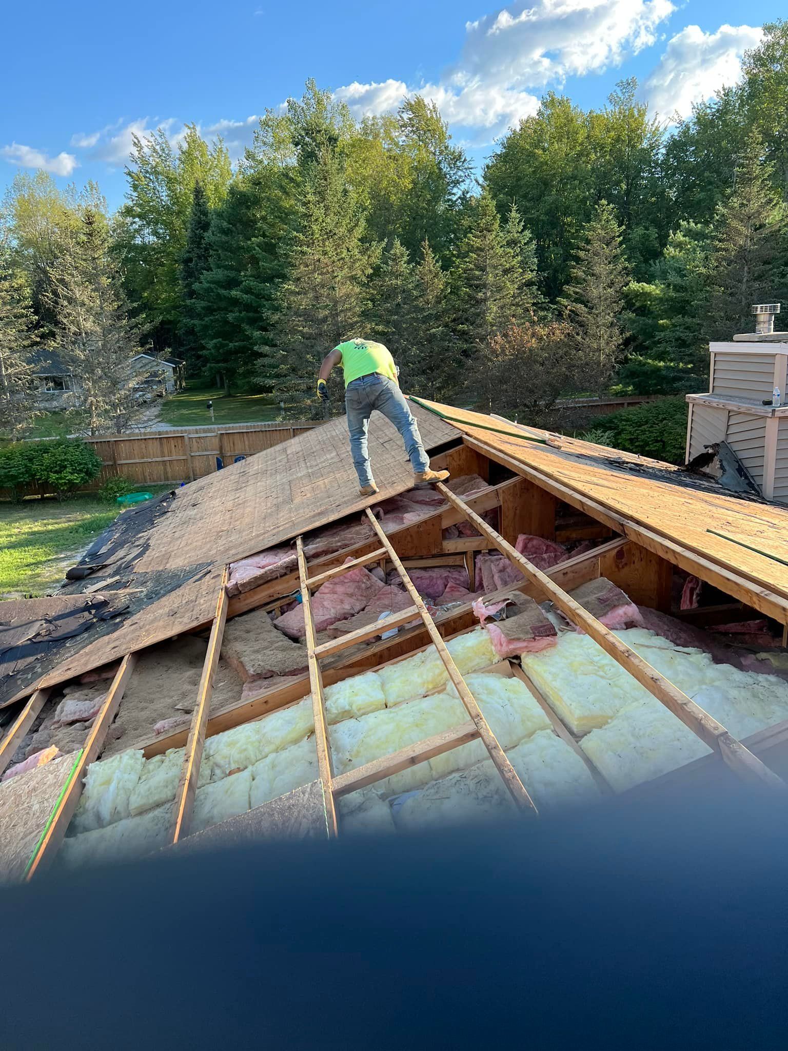 Roofer on a partially torn-off roof. The sky is blue with clouds, surrounded by trees.
