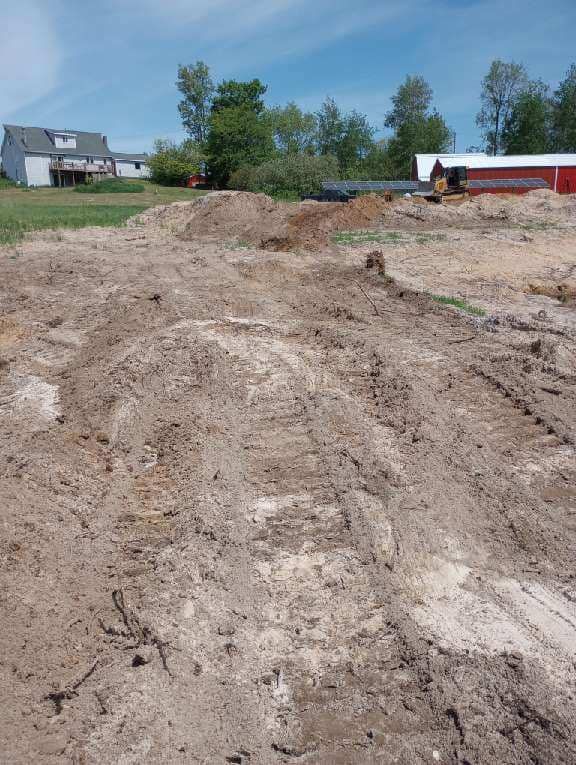 Dirt field with tractor tracks; house and red barn in the background under a blue sky.