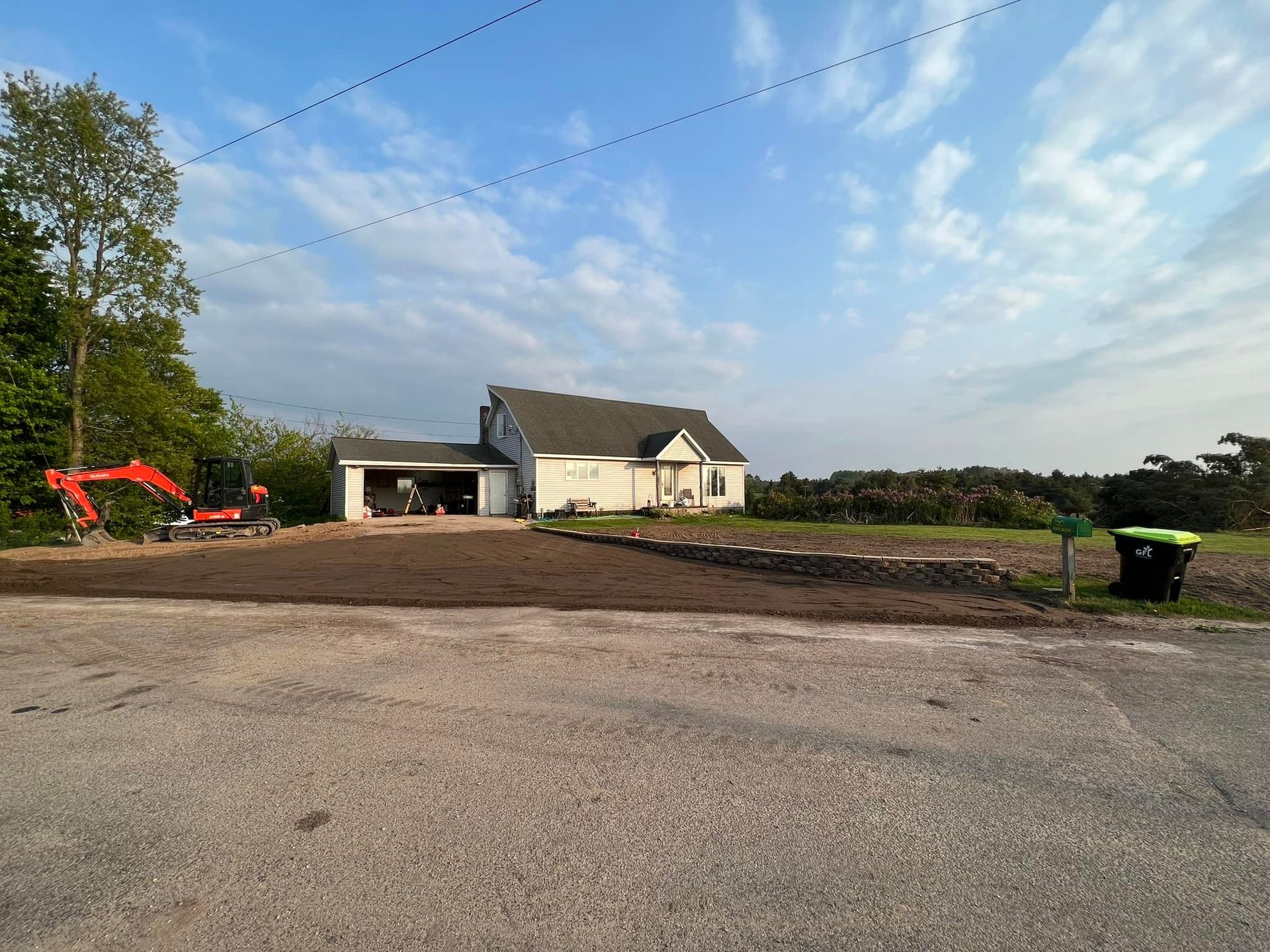 House with detached garage, freshly tilled yard, and small excavator. Cloudy sky.