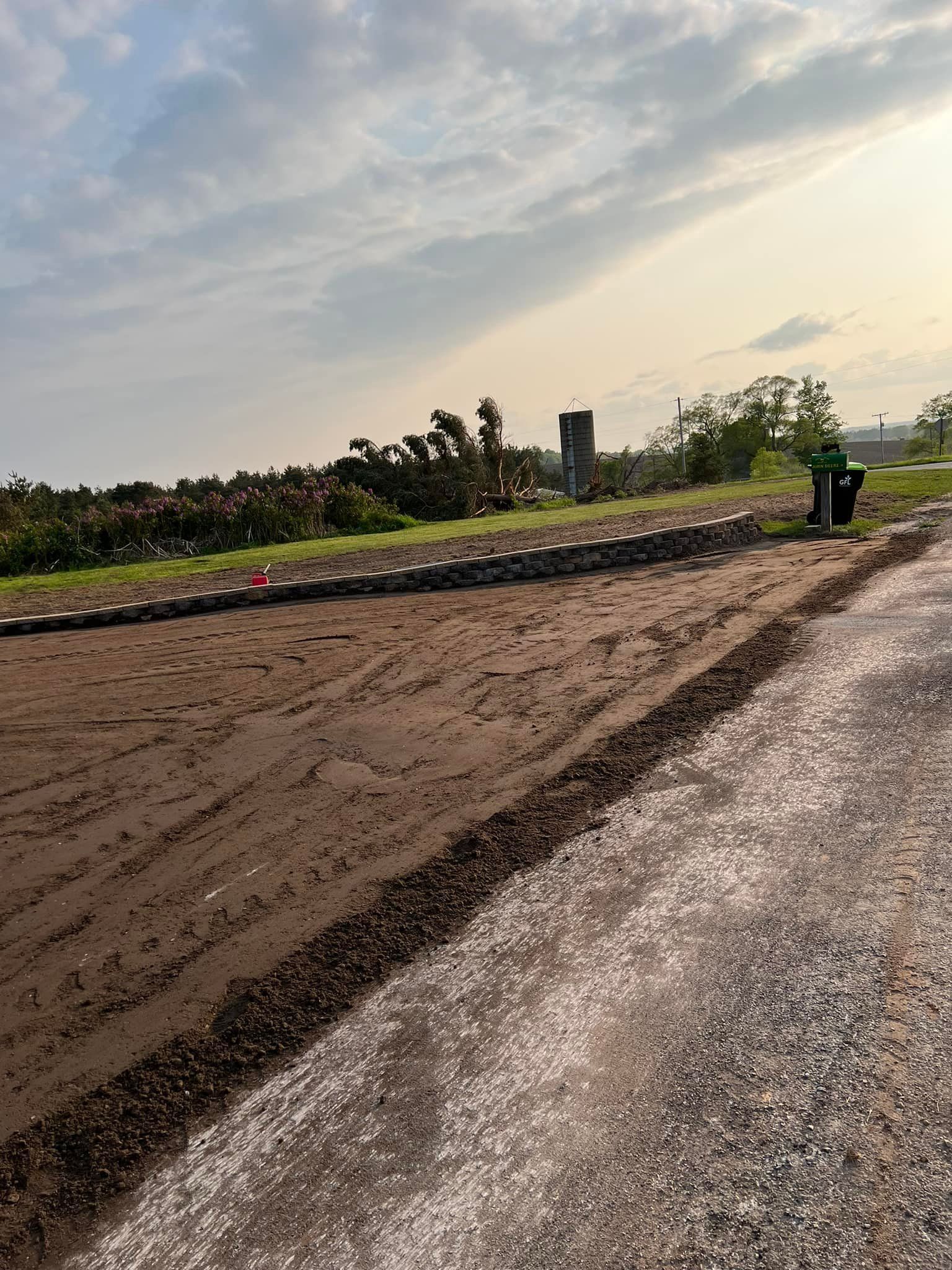 Dirt patch beside gravel road with a row of small white flags in a field with trees.