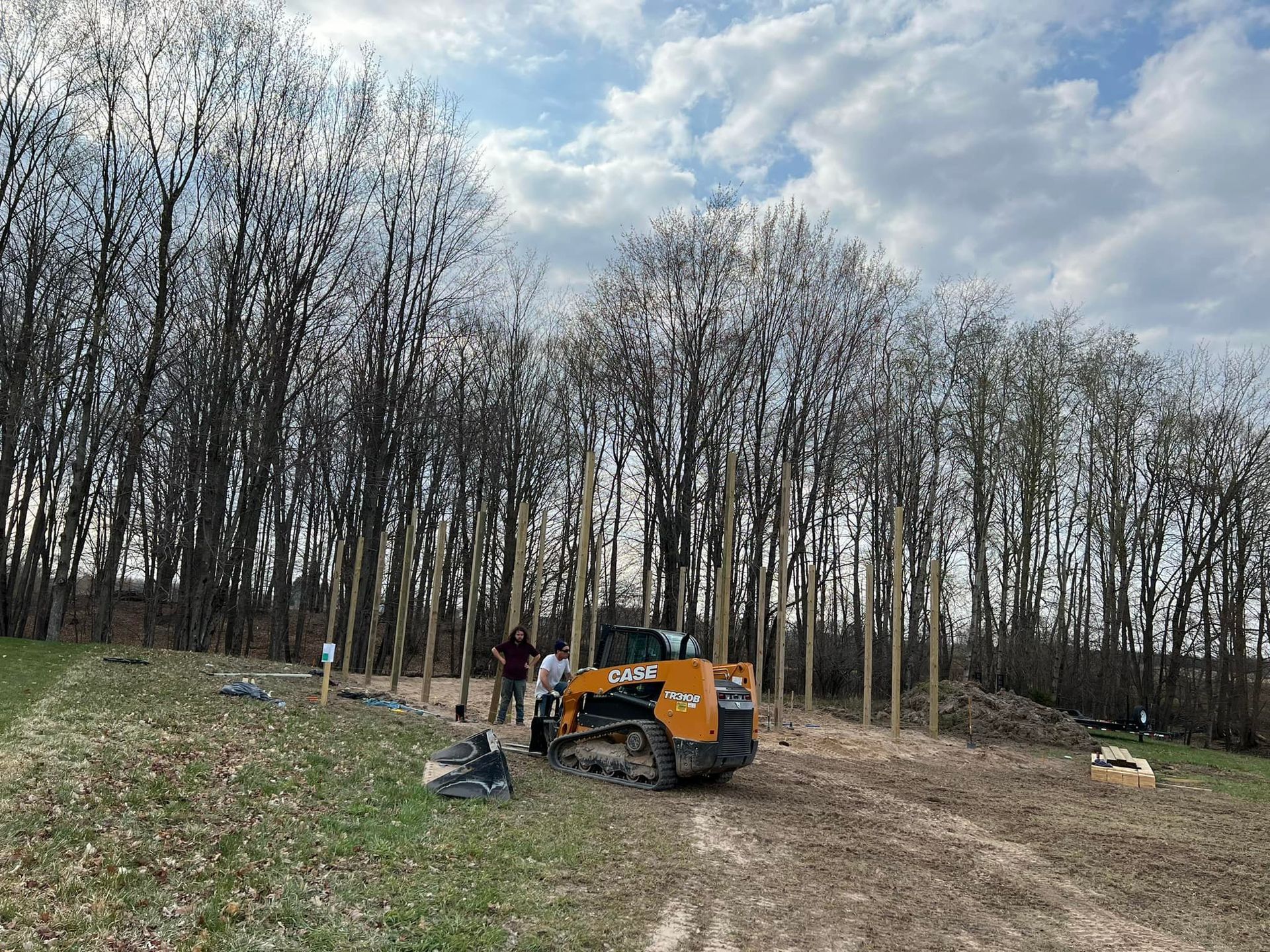 Construction site: Skid steer, workers, and posts set in the ground, among trees, under a cloudy sky.