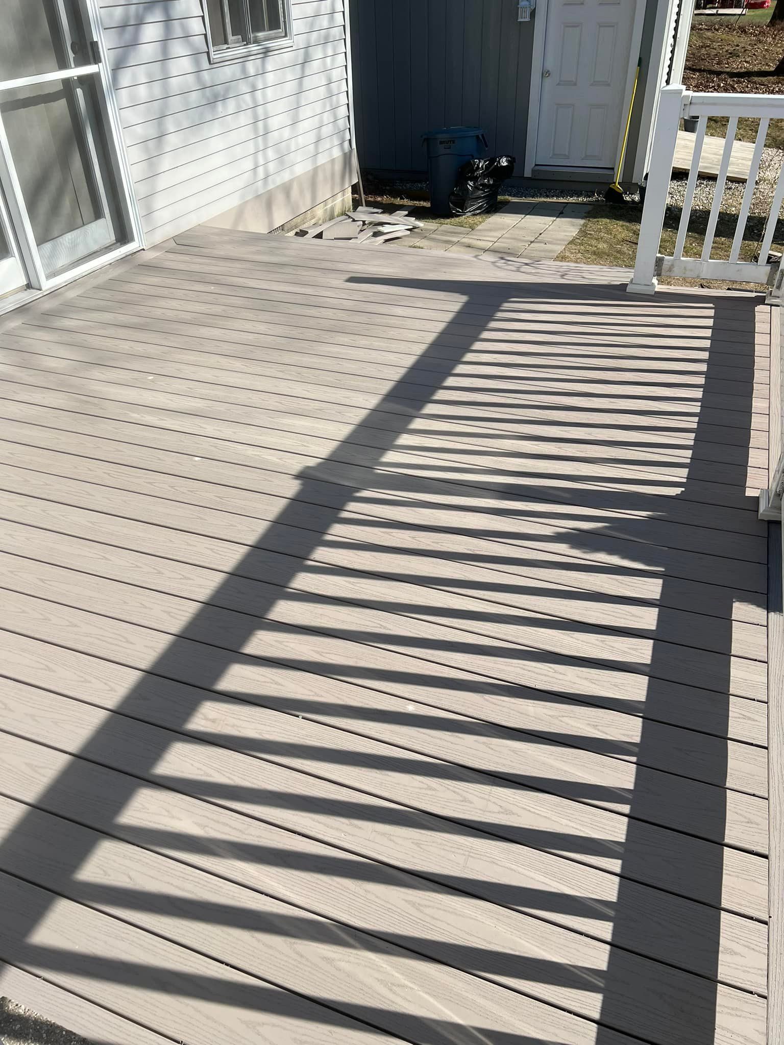 Deck with a shadow cast from a railing, next to a white house with a sliding glass door.