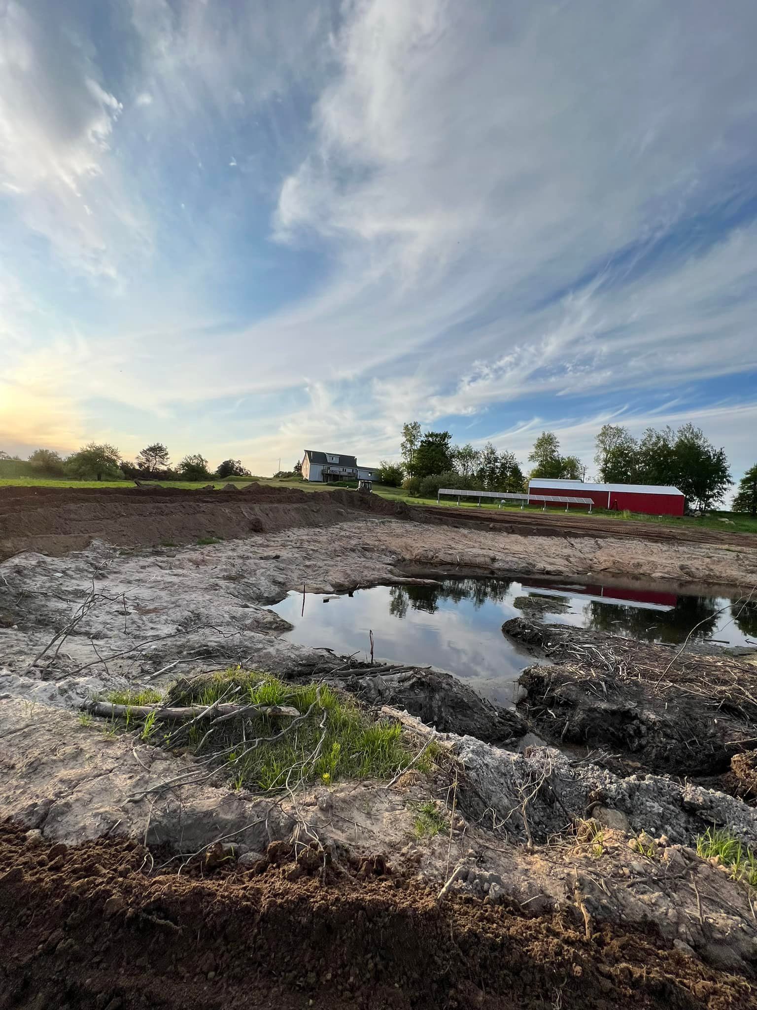 Puddle in a dirt field under a cloudy blue sky. Red barn and trees in the distance.