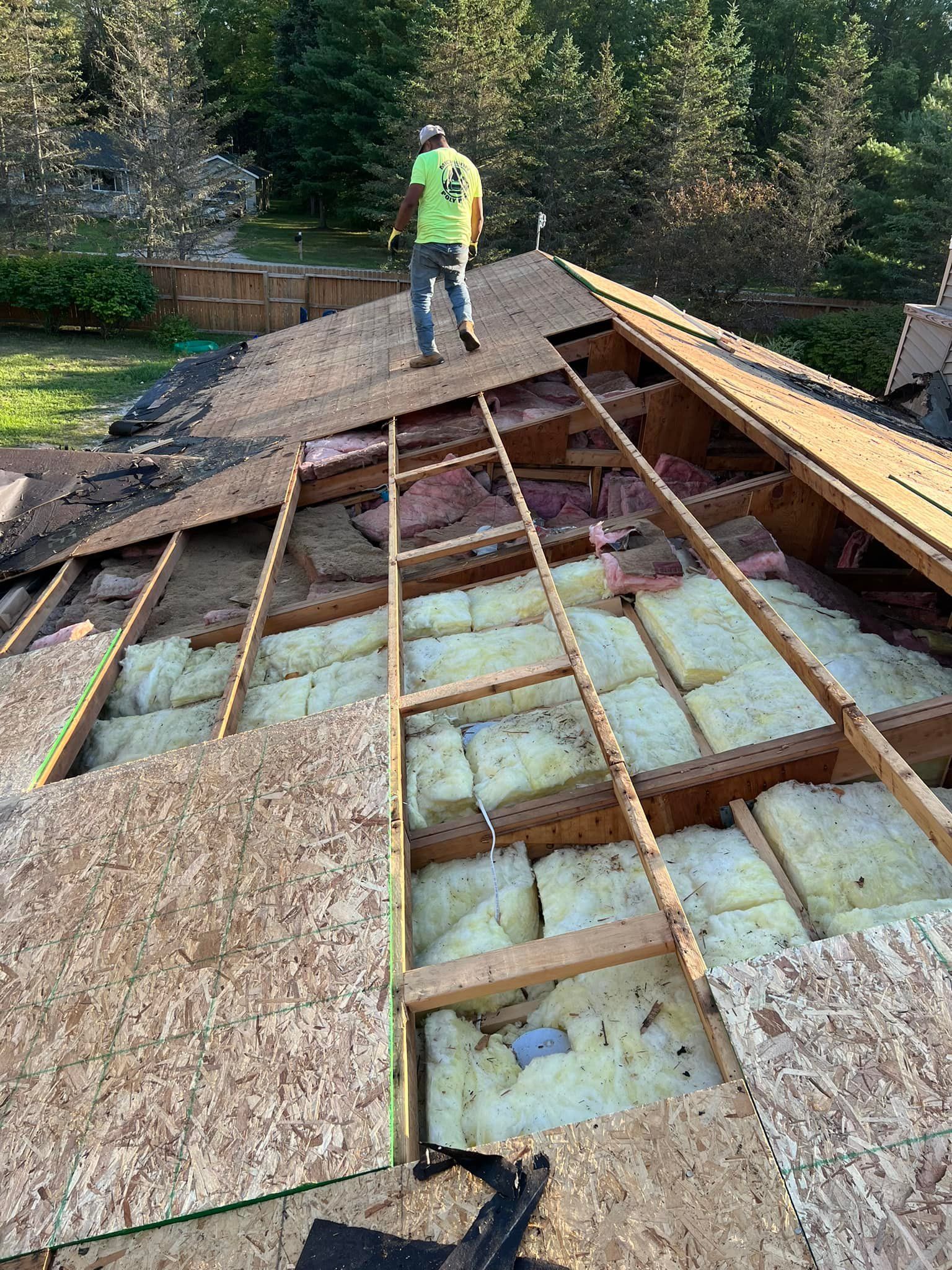 Person on a partially deconstructed roof, exposing insulation. Green shirt, jeans, and trees in the background.