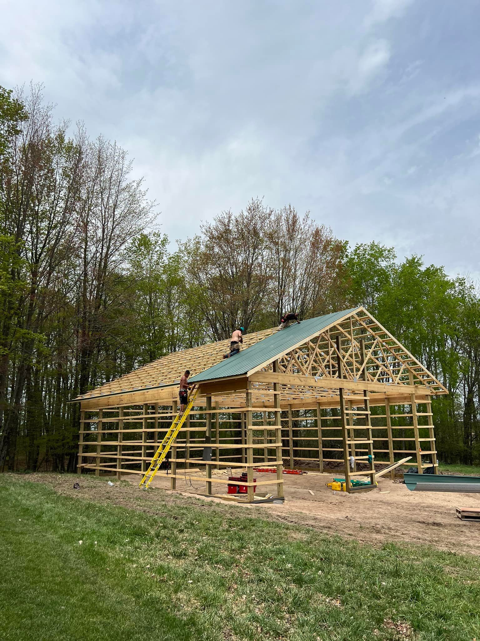 Construction of a wooden barn frame in a grassy field, workers on the roof, trees in background, overcast sky.