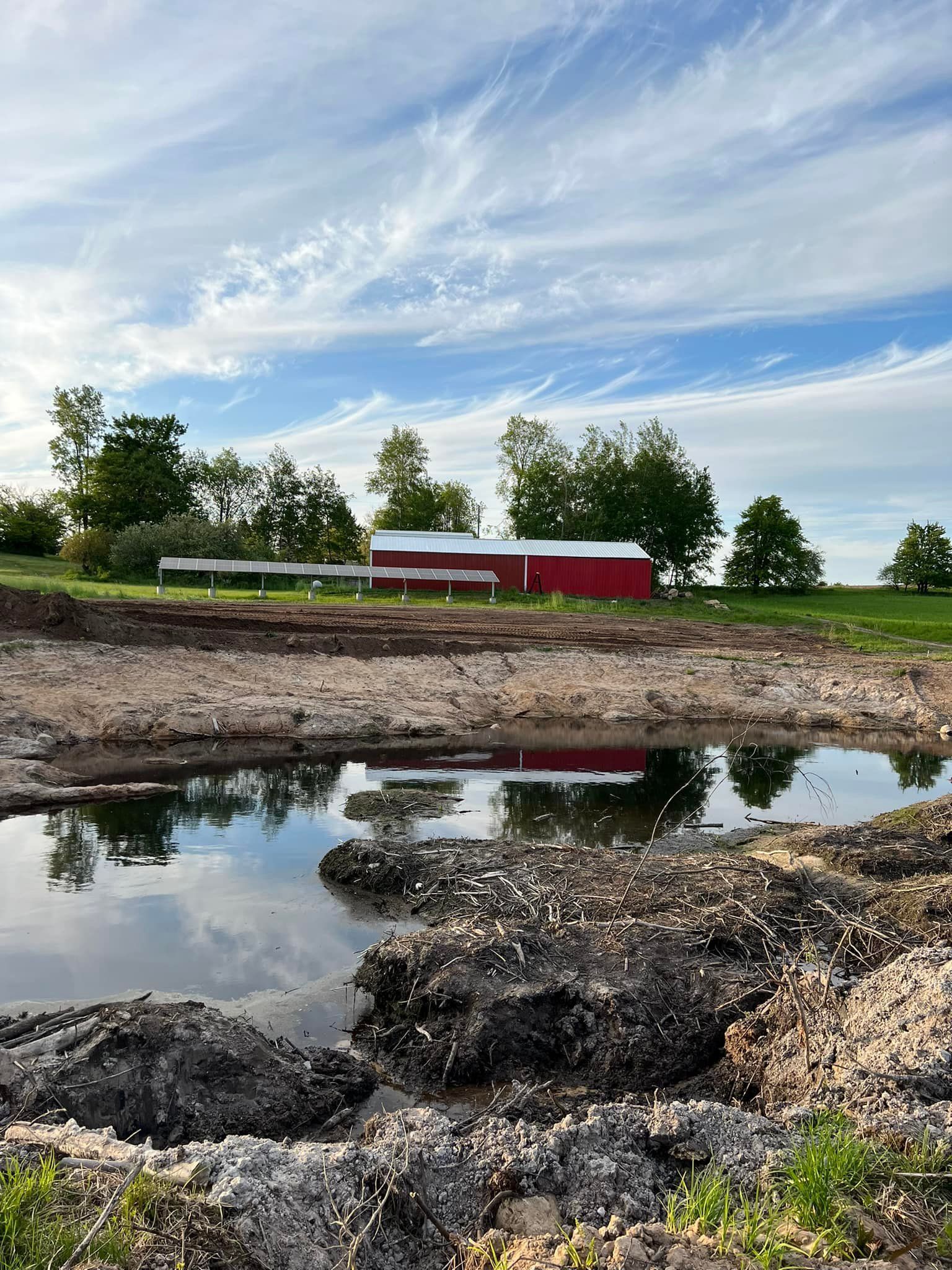 A red barn and trees reflected in a muddy pond under a blue sky with streaked clouds.