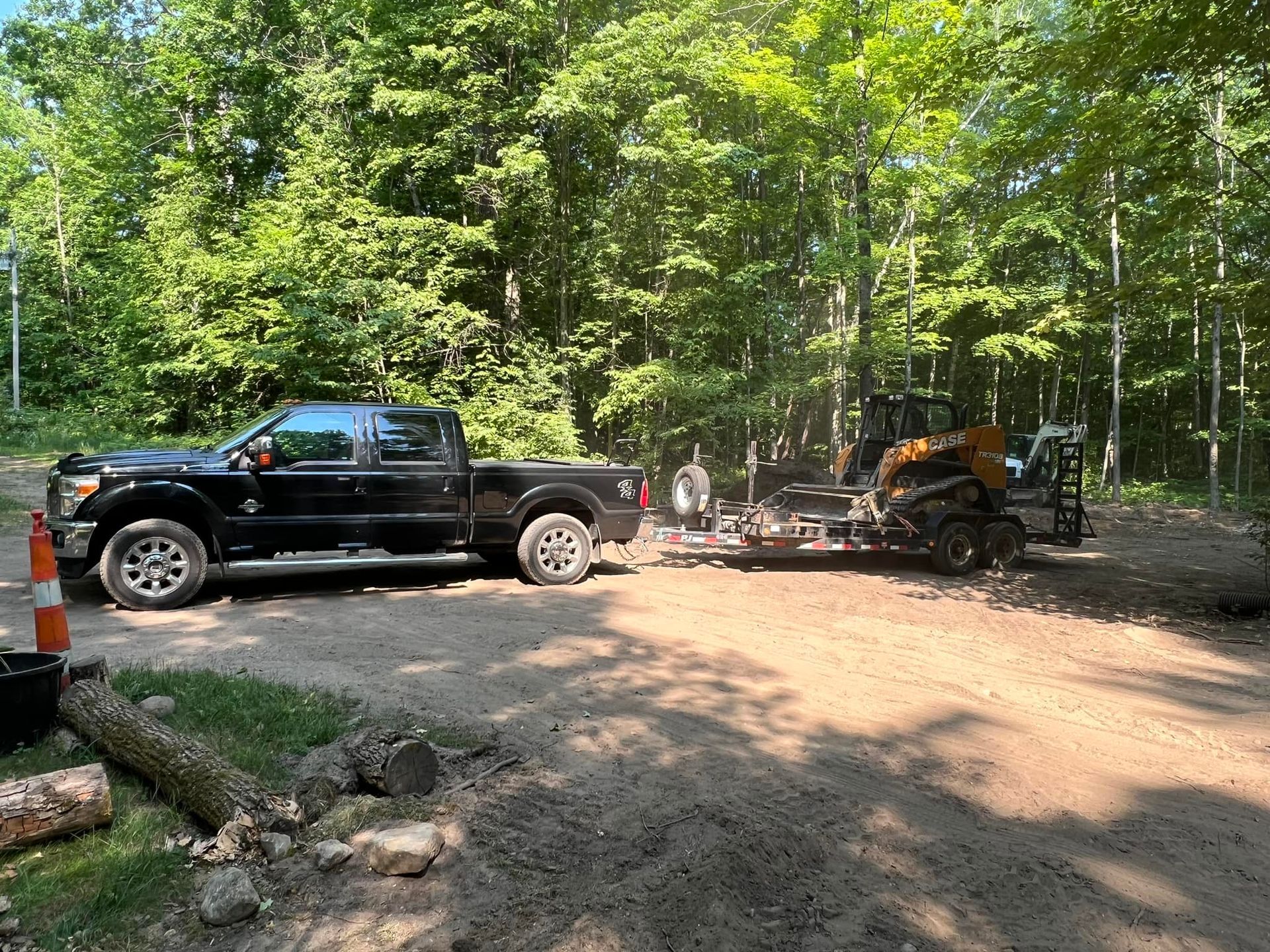 Black pickup truck towing a trailer with a small excavator on a gravel road in a wooded area.