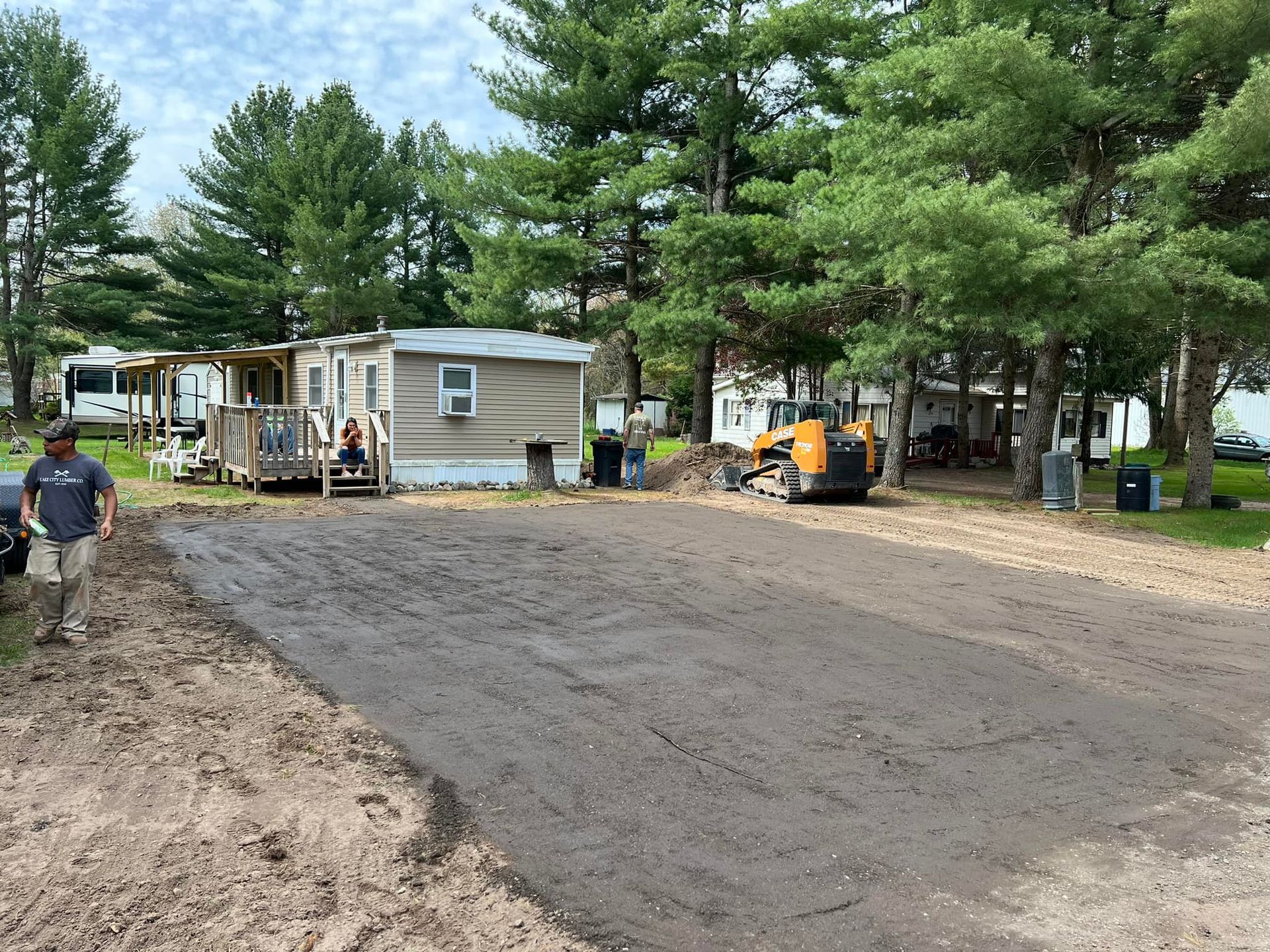 A man stands in front of a mobile home with construction equipment nearby. Gravel and dirt cover the area.