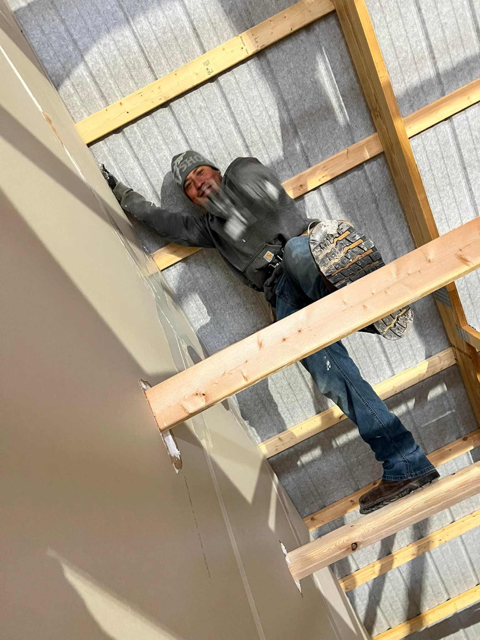 Construction worker on roof frame, reaching, wearing safety gear, grey and blue.