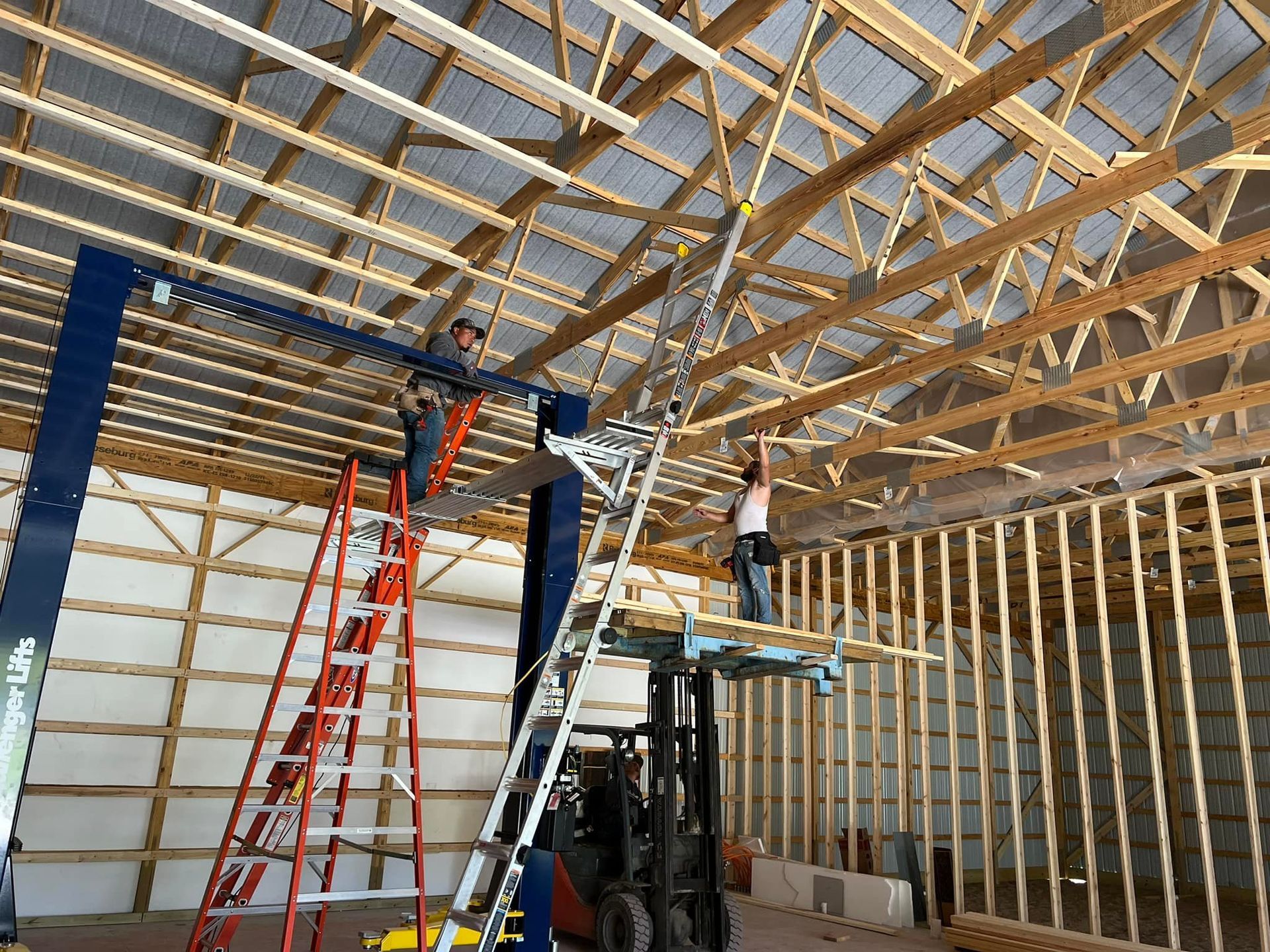 Construction workers installing lights in a barn; one on a ladder, two on lifts, wood beams and supports visible.